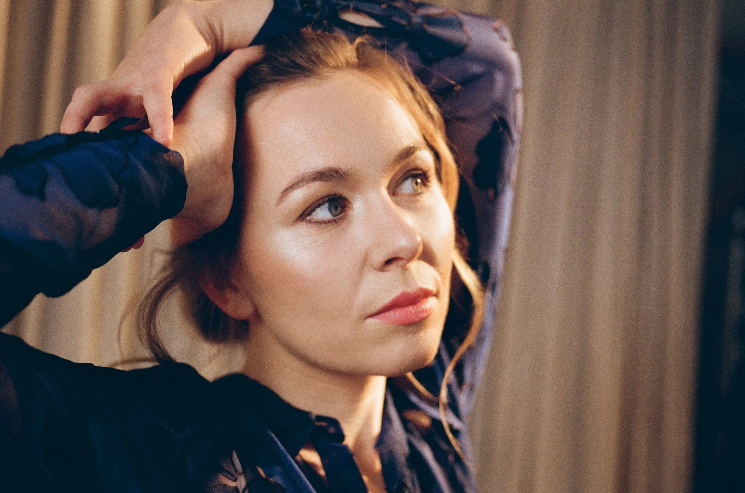 Elsa Dreisig opera singer with red hair and blue eyes gazes to the side, relaxing with her hands on her head. She wears a dark, sheer, floral-patterned blouse and sits against a background of beige curtains.