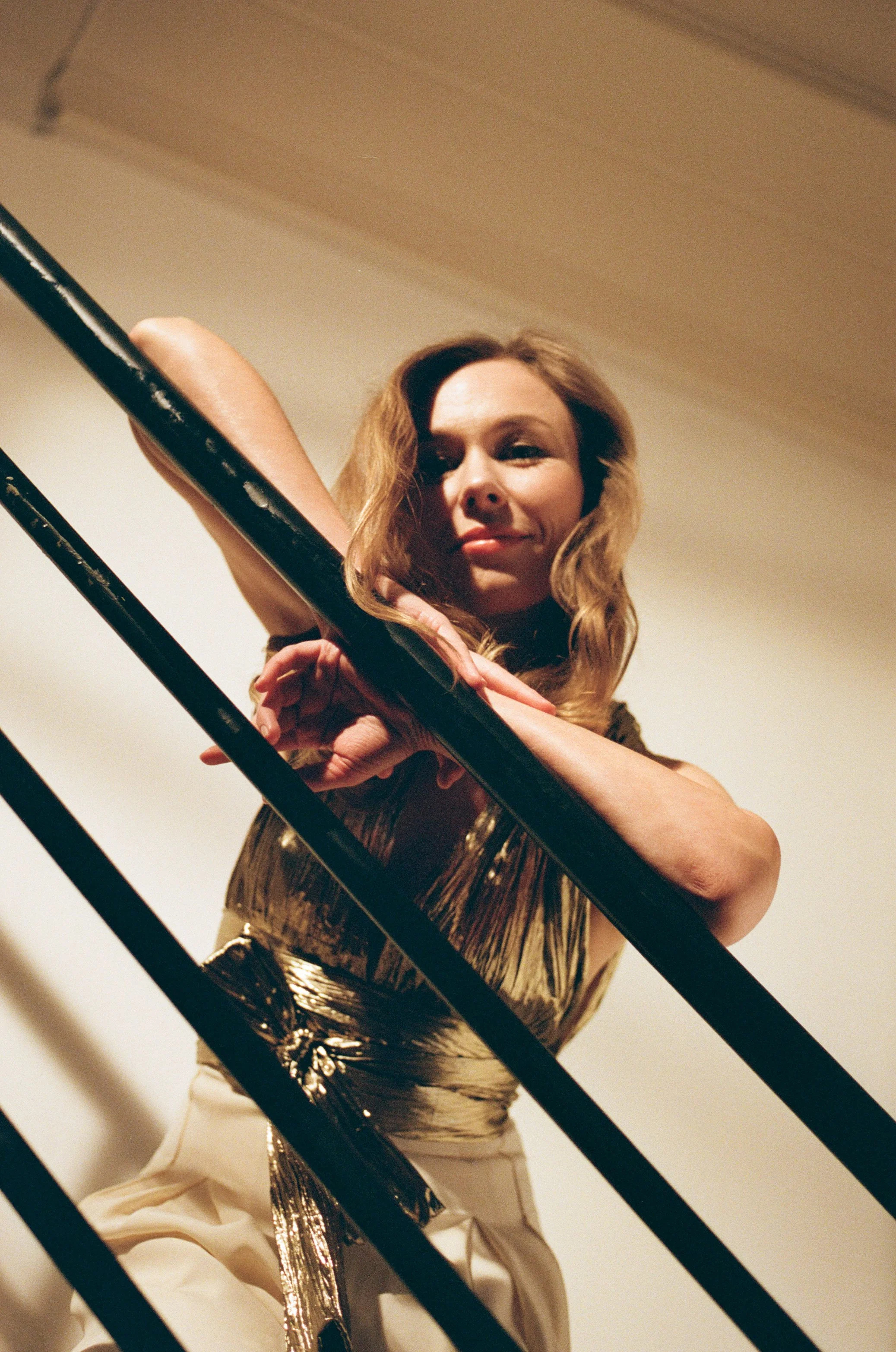 A woman with wavy hair leaning on black metal stairs railing, looking confidently at the camera.