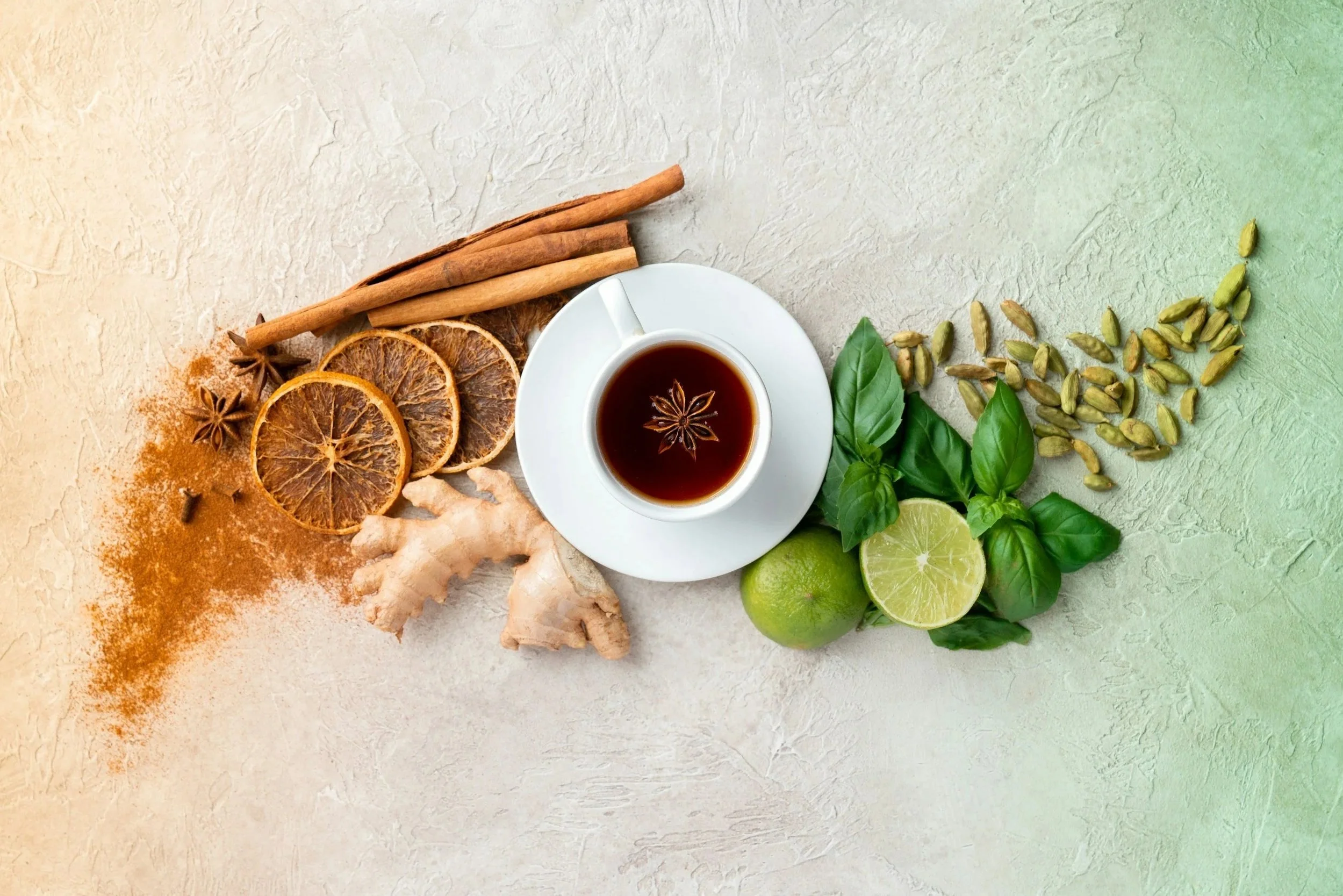 A top view of a cup of black tea on a white saucer surrounded by dried orange slices, cinnamon sticks, star anise, fresh ginger, a lime, basil leaves, and green cardamom pods on a light textured surface.