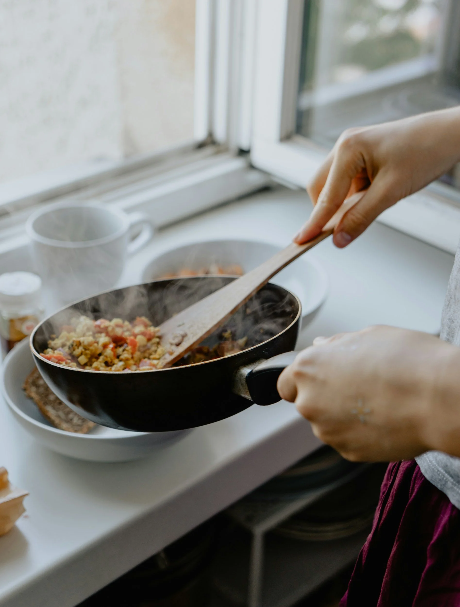 Person cooking scrambled eggs in a black frying pan on a white kitchen counter near a window.
