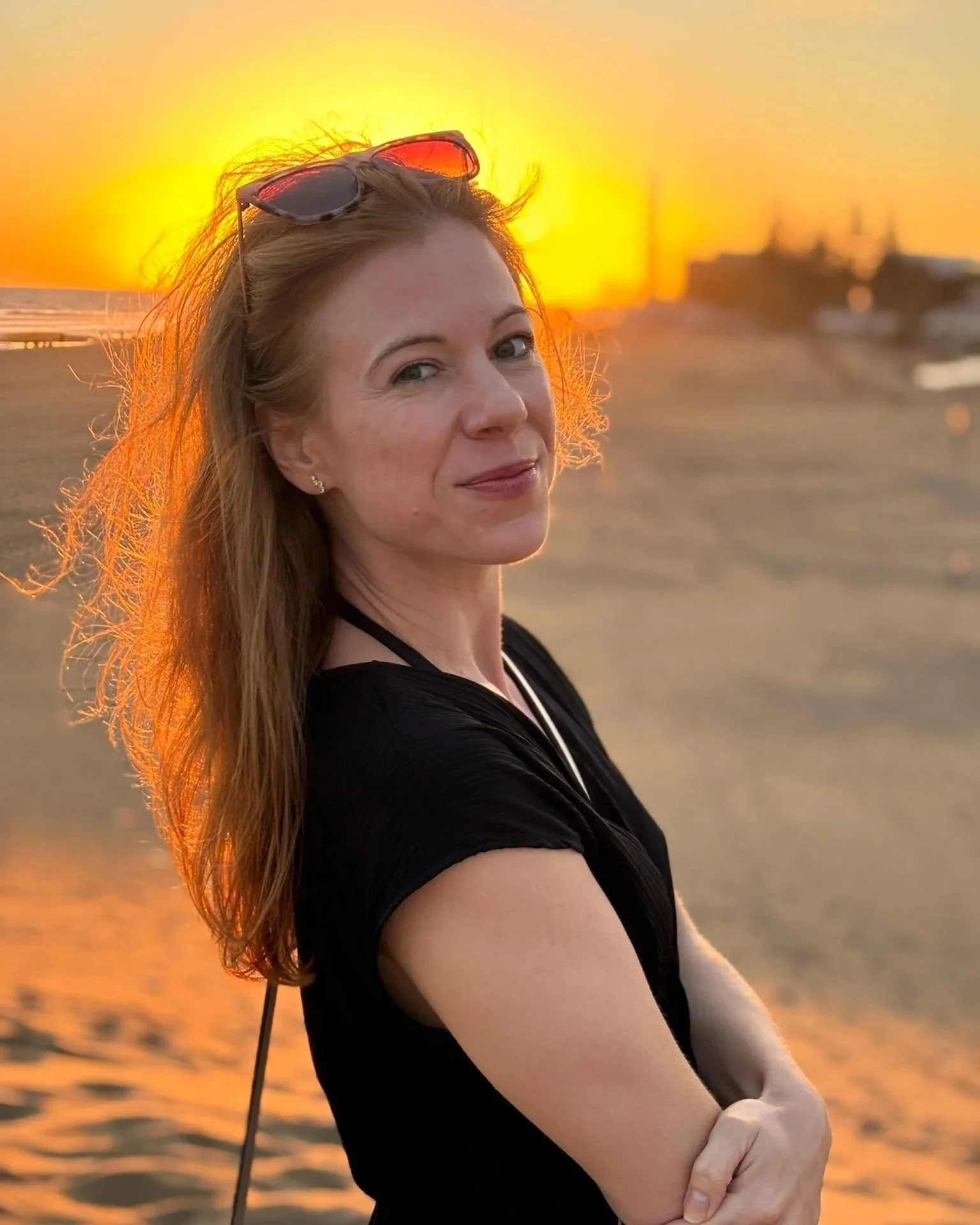 A woman with long red hair, wearing sunglasses on her head and a black top, stands by the water during sunset, with the sun setting behind her and creating a golden glow.