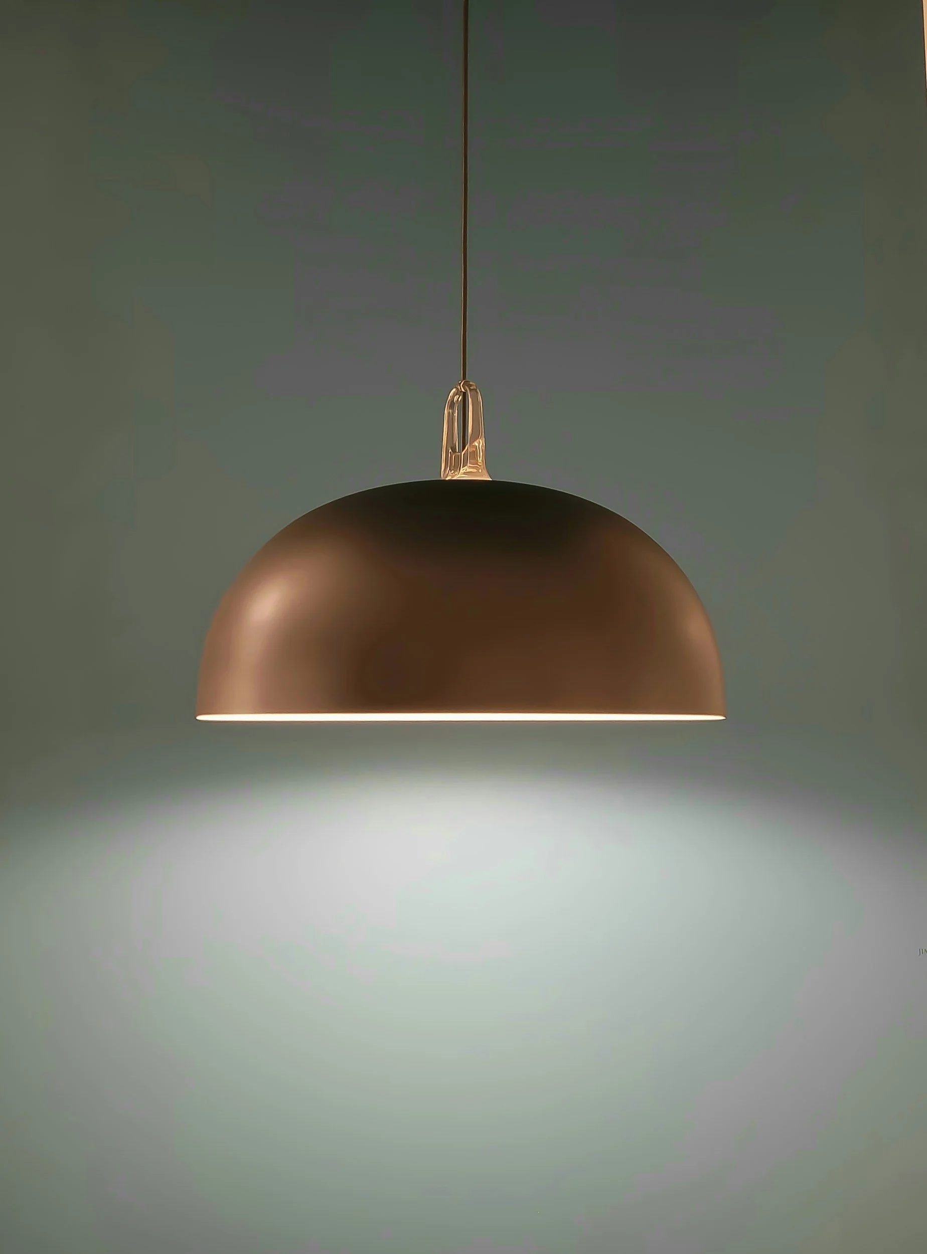 Lámpara moderna suspendida, de color bronce, iluminando desde el techo contra fondo gris.