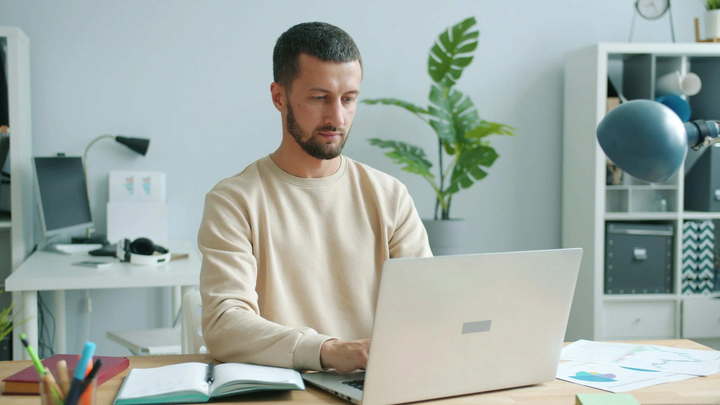 Hombre trabajando en una sala de oficina con una laptop en un escritorio de madera, con libros, papeles y artículos de oficina, y una planta en el fondo.
