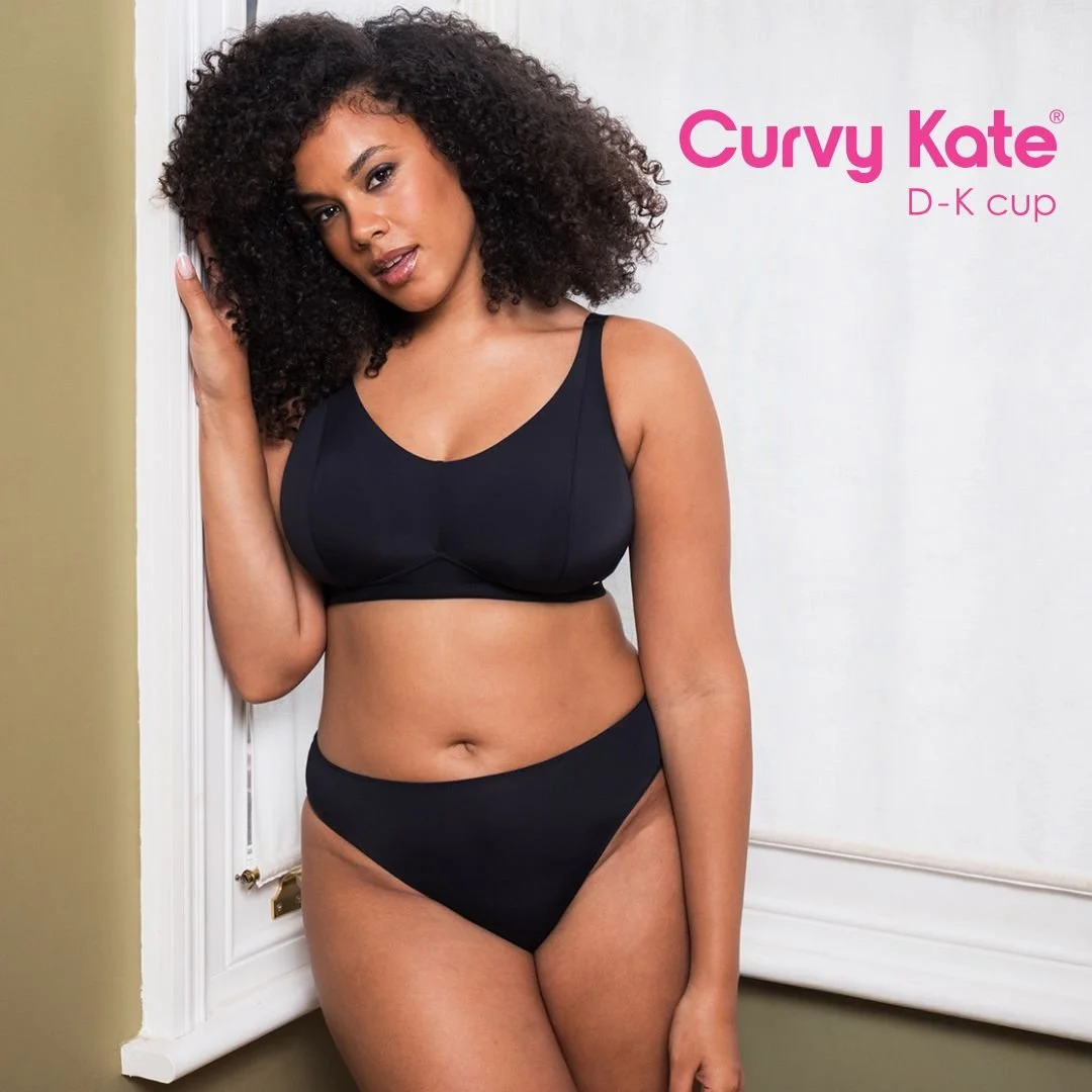 A woman with curly hair wearing a black bra and matching underwear, standing next to a white wall with a small section of green wall visible, advertising Curvy Kate D-K cup bras.