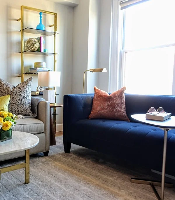 A living room corner with a beige sofa, a dark blue sofa, a white coffee table with a bouquet of yellow flowers, a gold and glass shelving unit with decorative objects, a table lamp, a floor lamp, and a window with sunlight.
