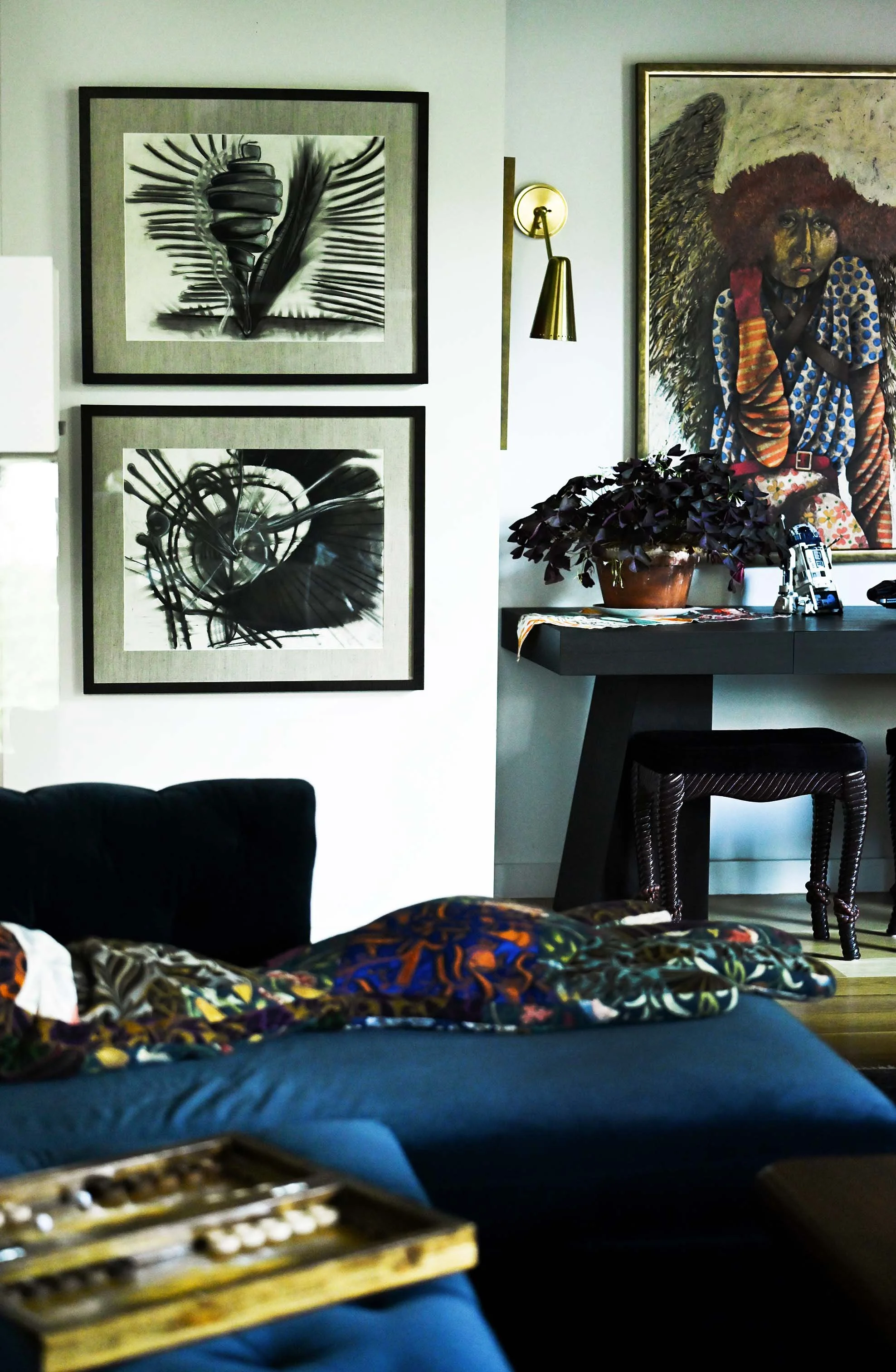Living room with black and white abstract art on the wall, a dark side table with a potted plant, a large colorful artwork of an African woman, and a partially visible sofa with colorful fabric on top.