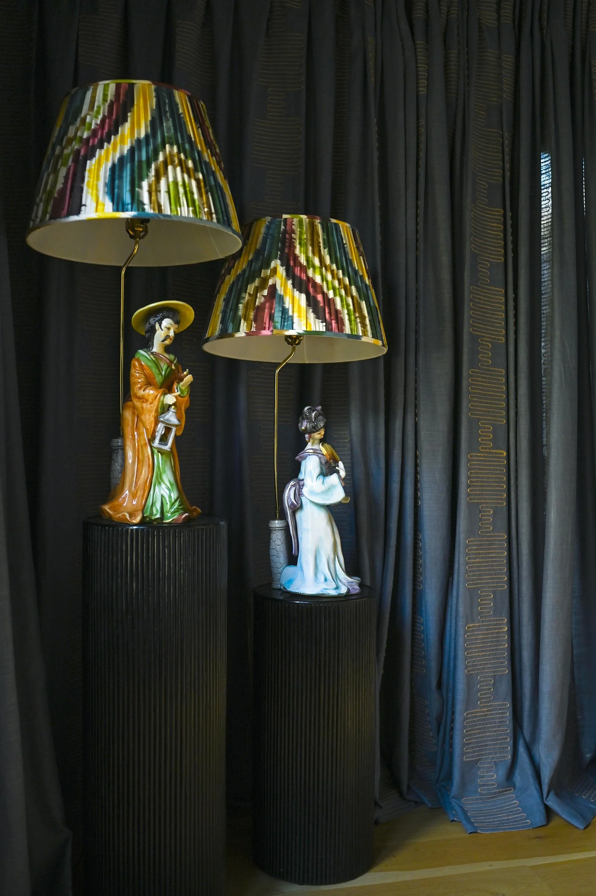 Two ceramic figurines of women dressed in traditional Asian attire, each used as a lamp base with colorful patterned lampshades, positioned on black pedestal stands in front of dark curtains.