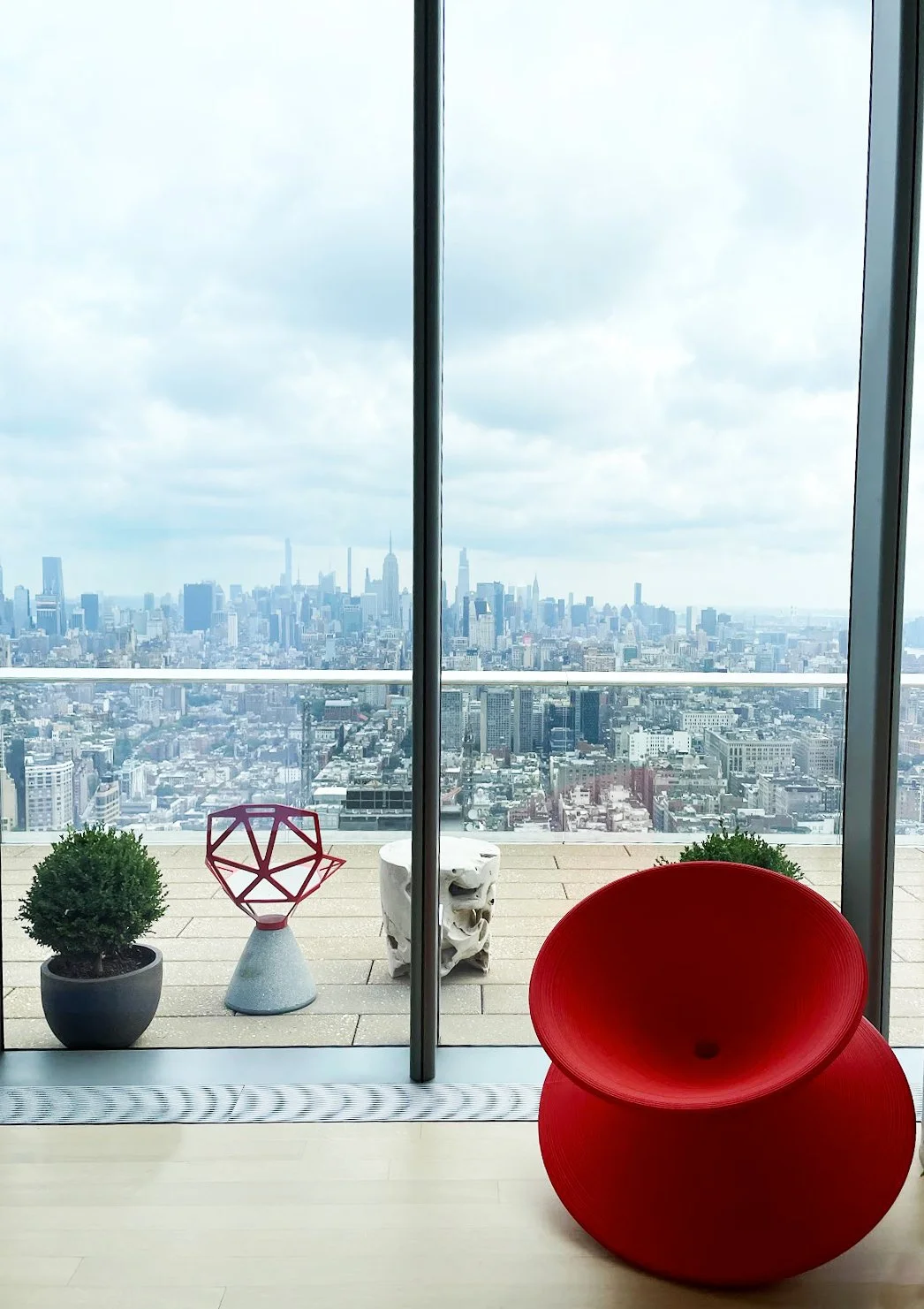 Interior of a high-rise building with floor-to-ceiling windows showing a city skyline, potted plants, and modern furniture including a red chair and a small pink geometric sculpture.
