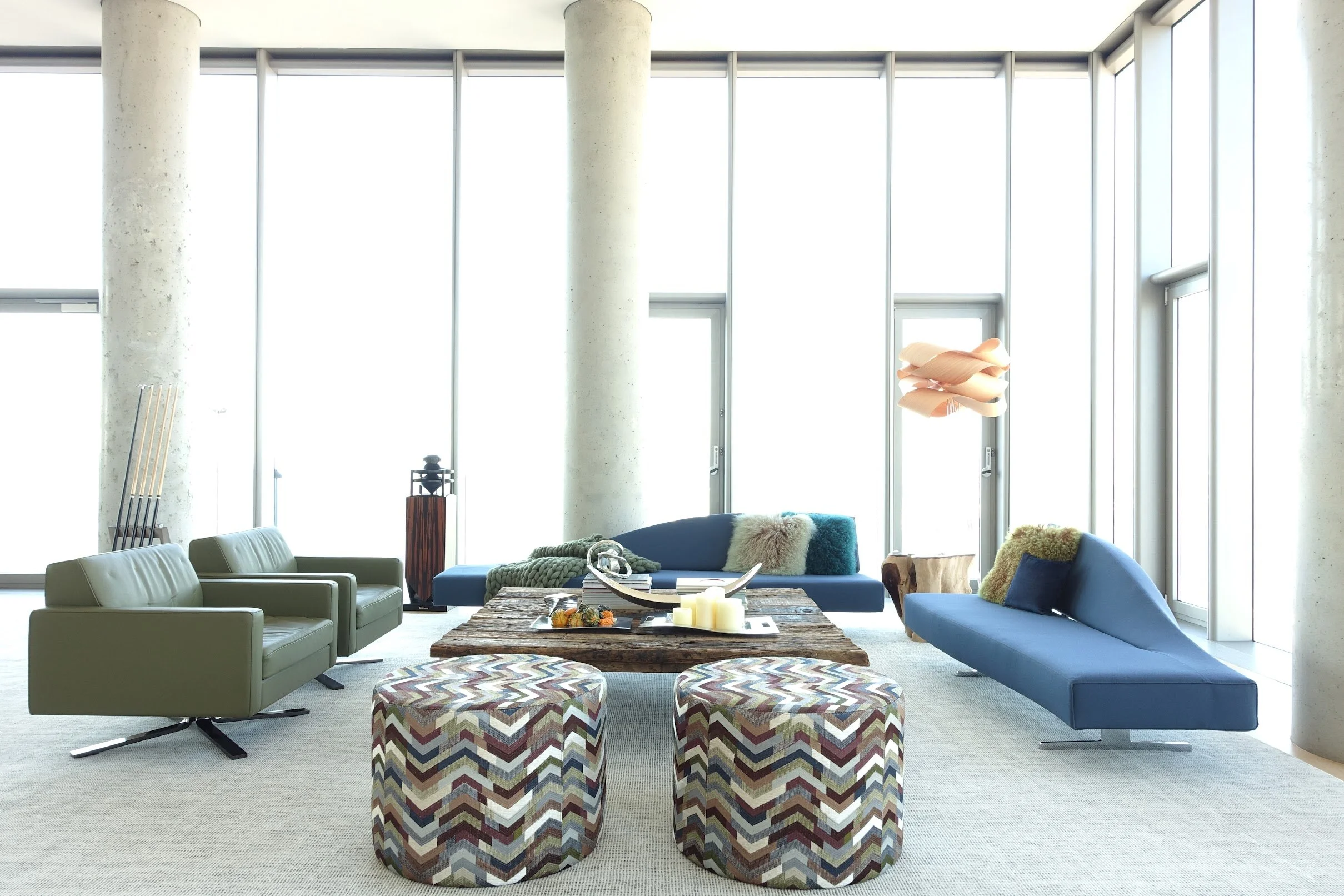 Modern living room with large windows, including blue and green sofas, a rustic coffee table, patterned ottomans, and decorative objects.