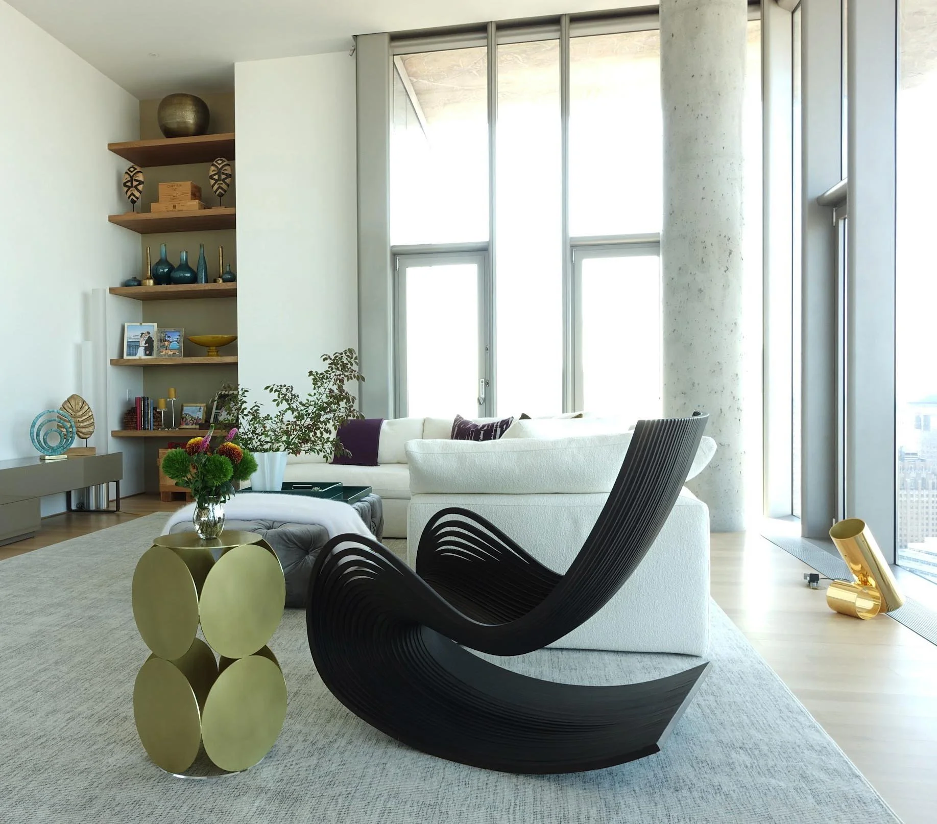 Modern living room with large windows, white sofa with purple pillows, black sculptural lounge chair, gold side table with a floral arrangement, and built-in wall shelves with decorative items.