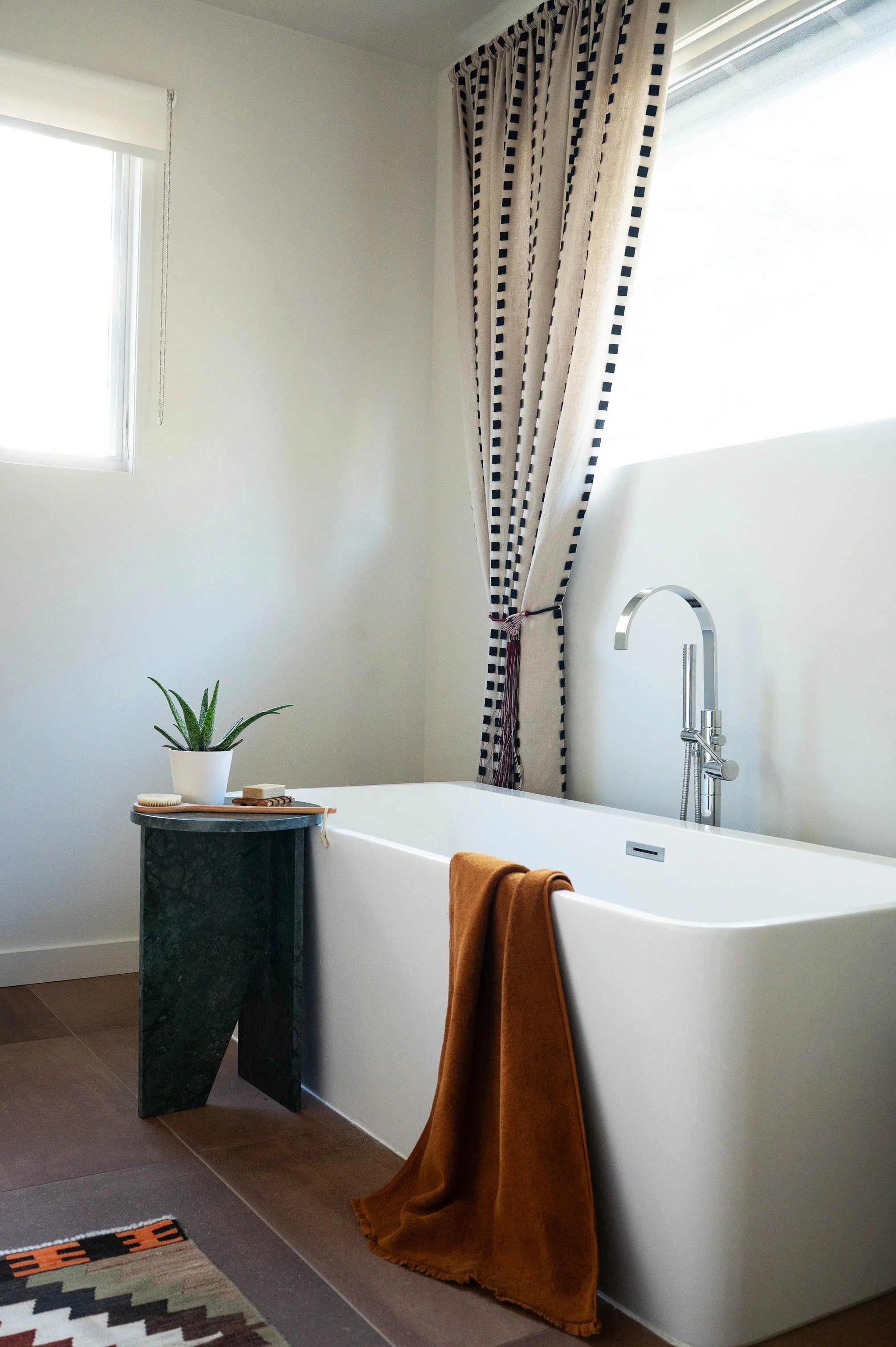 Modern bathroom with a white bathtub, a green side table, a potted plant, a rust-colored towel, and black and white patterned curtains next to a large window.