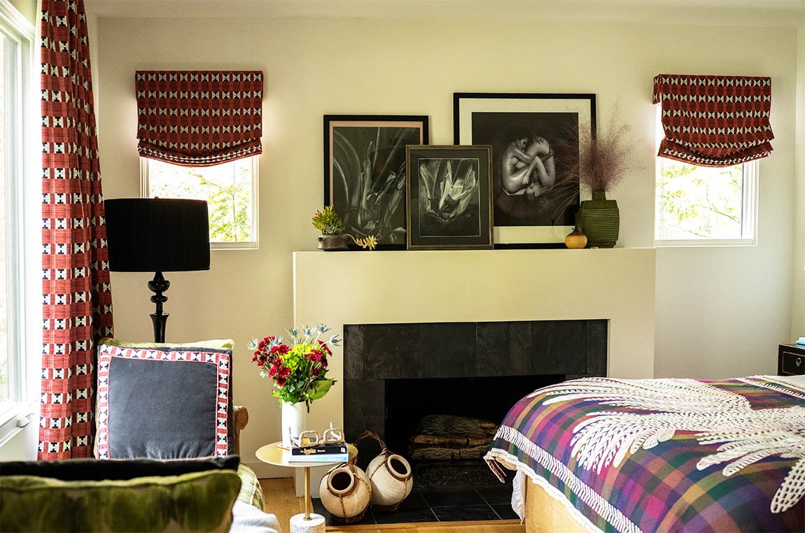 A cozy bedroom with a fireplace, decorated with framed artwork, vases, and plants. There are two small windows with red patterned curtains, a black lampshade, a bedside table with a vase of flowers, and part of the bed with a plaid blanket.
