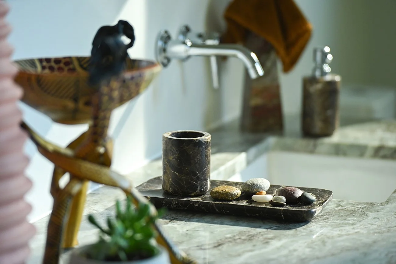 A bathroom countertop with a black stone cup, various colorful stones on a black marble tray, soap dispensers, a brown towel, and decorative items.