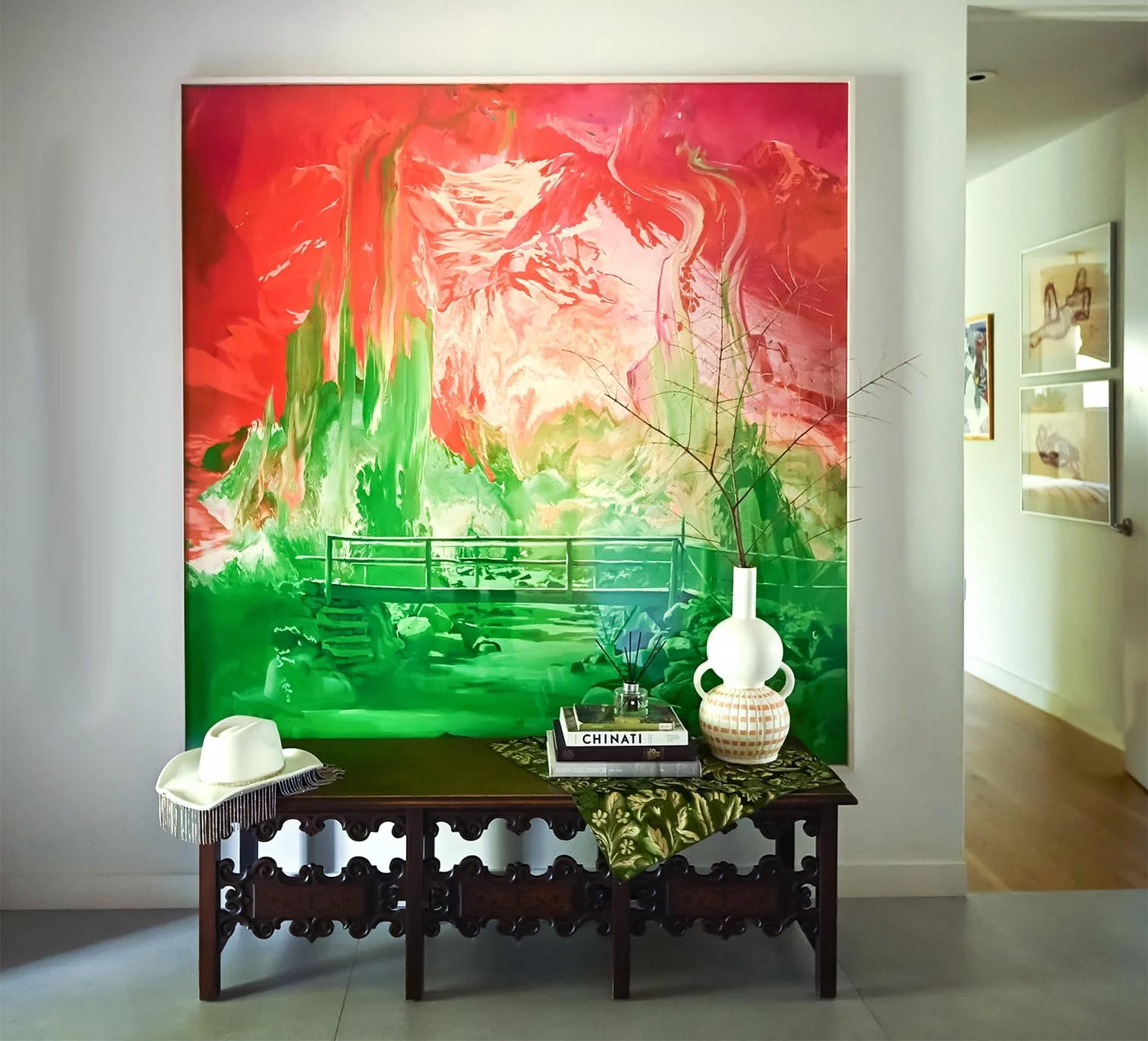 Decorative table with a book titled 'CHINATI', a white vase with green branches, a white hat with fringe, and a patterned cloth, in front of a large, colorful abstract mural with reds, pinks, and greens.