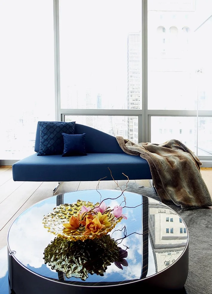 Modern living room with a blue sofa, cushions, a blanket, and a glass-top coffee table with a floral arrangement, large windows showing city buildings.