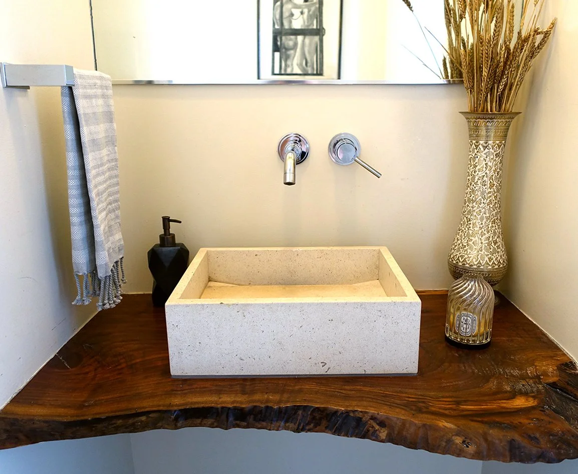 A bathroom sink with a rectangular stone basin mounted on a wooden counter, a black soap dispenser to the left, a mirror above, and a decorative vase with dried grasses to the right.