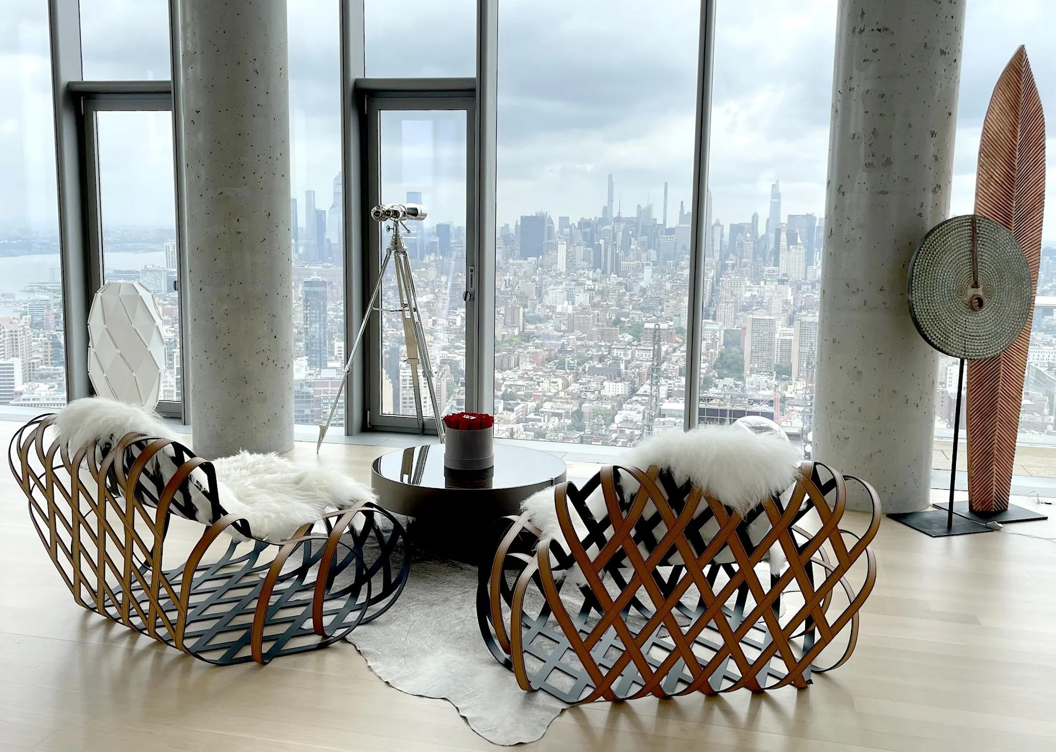 Modern living room with floor-to-ceiling windows offering a city view, two stylish curved wooden chairs with white cushions, a black coffee table with a red flower arrangement, a telescope near the windows, a large decorative surfboard, and a tall ro