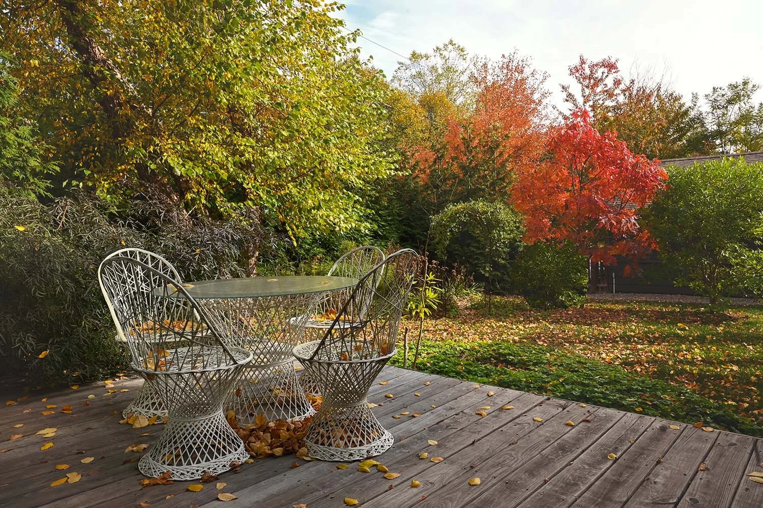 A wooden deck with a round glass top table and four metal wire chairs, fallen leaves on the deck, surrounded by green and red autumn trees.