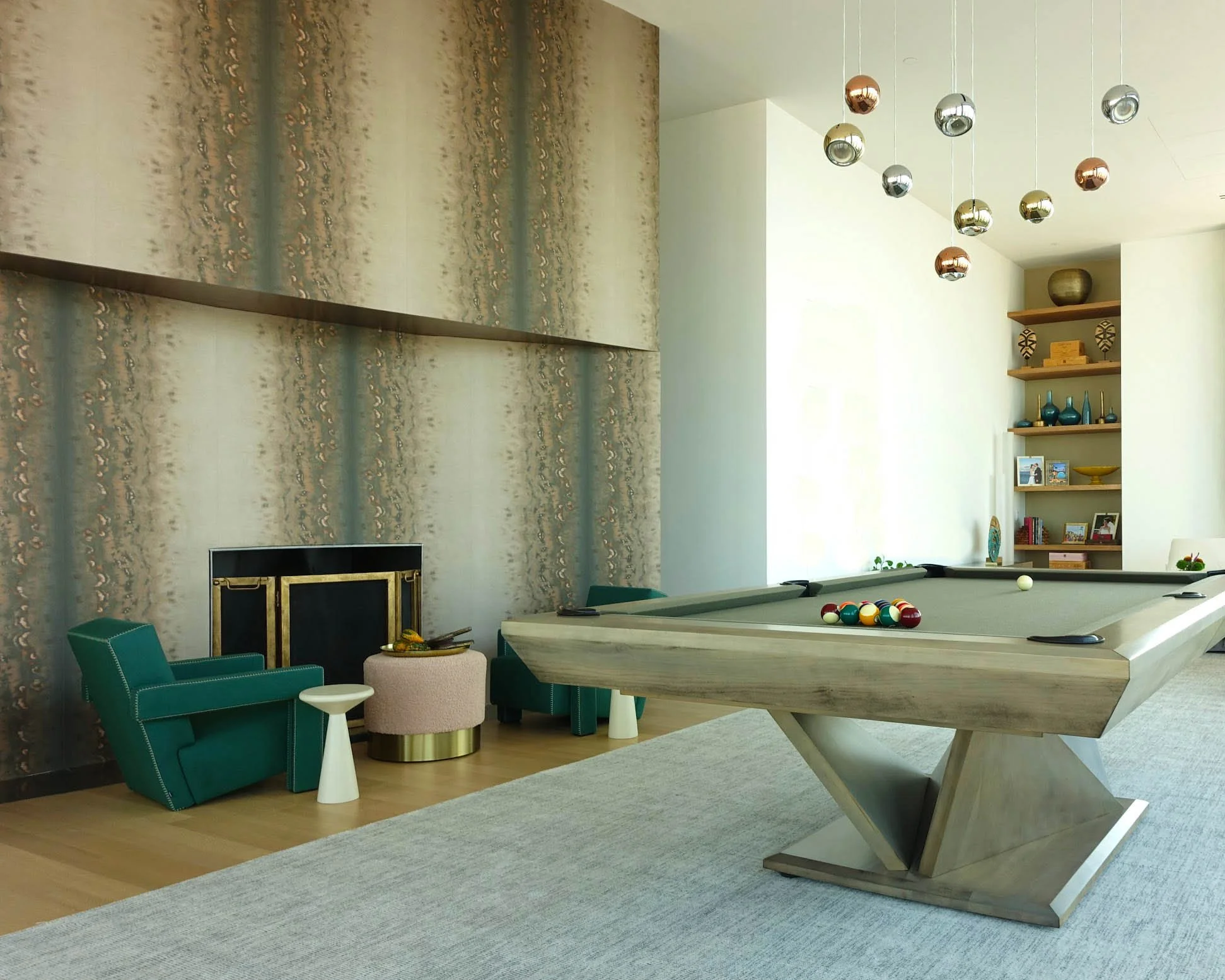 Living room with pool table, green armchair, beige ottoman, fireplace, corner shelves with decorative items, and hanging spherical light fixtures.