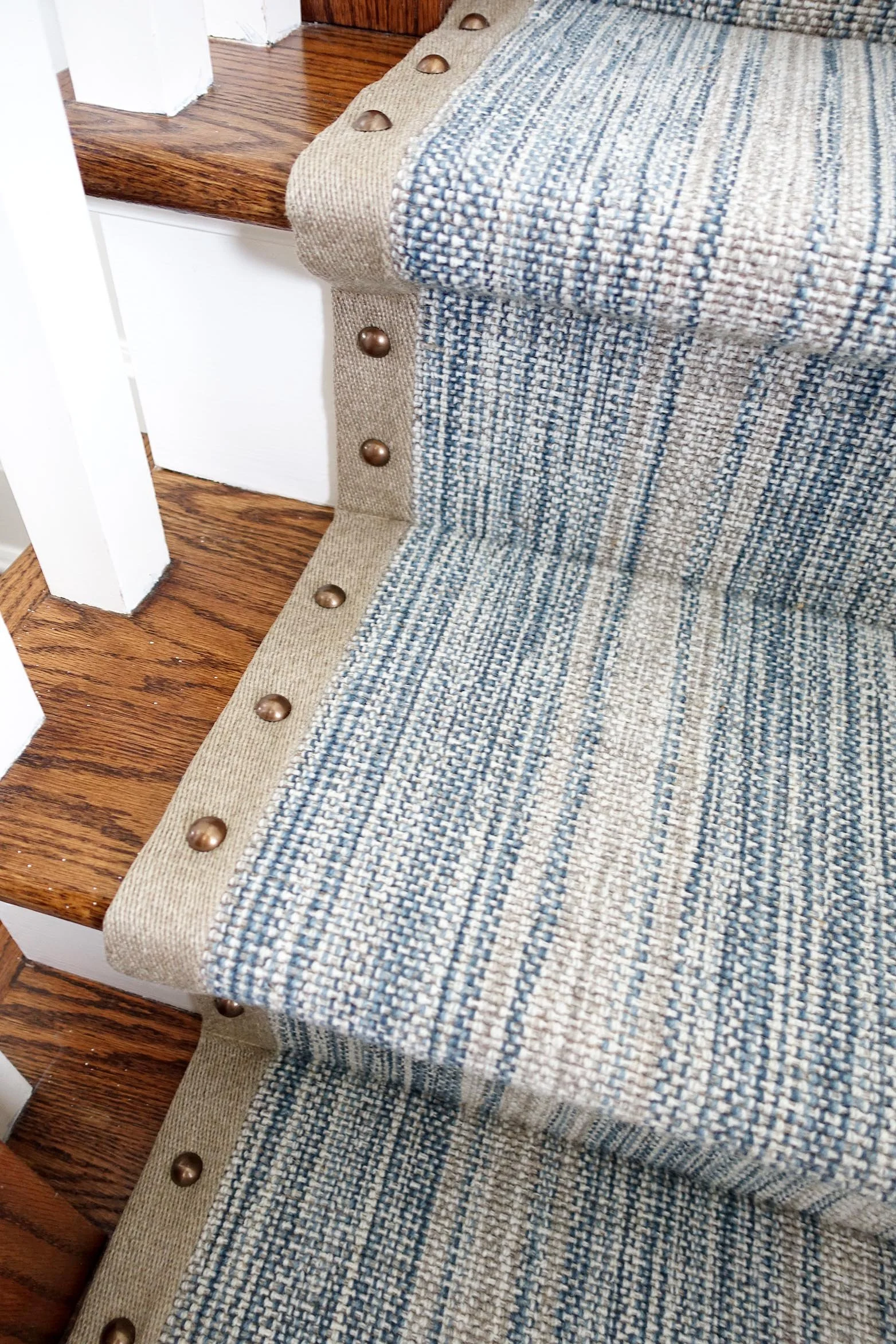 Close-up of a woven fabric staircase with blue, beige, and white stripes, with brass decorative studs along the edge, next to a wooden raised platform and white railing.