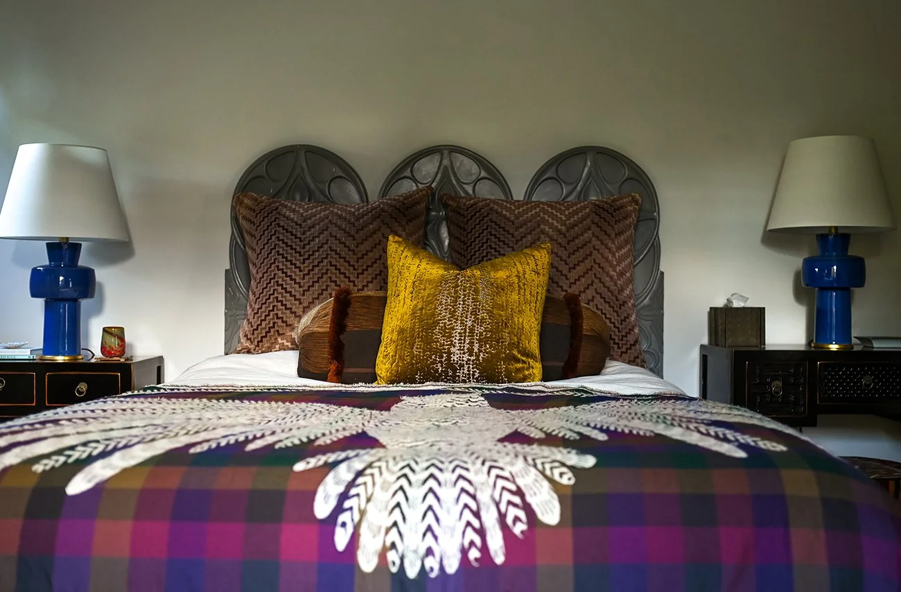 A neatly made bed with three large zigzag-patterned pillows, a vibrant yellow decorative pillow, and a bedspread featuring a plaid pattern with an embroidered white fern design. On each side of the bed are nightstands with matching blue lamps and sma