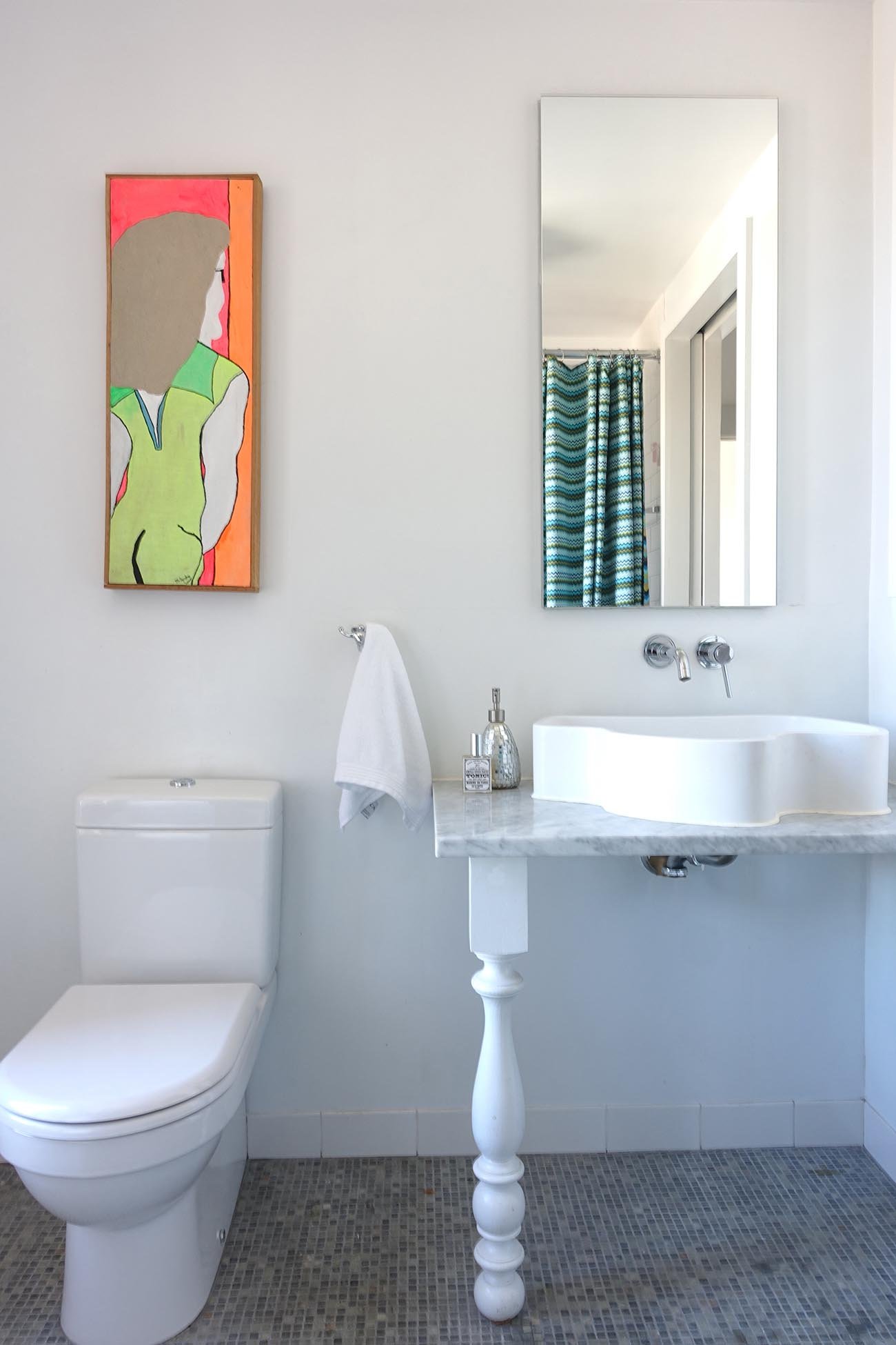 A modern bathroom with a white toilet, a marble countertop with unique sink, a mirror above the sink, a white towel hanging on the wall, a small decorative bottle on the countertop, a framed abstract art piece of a woman, and a mirror reflecting a do