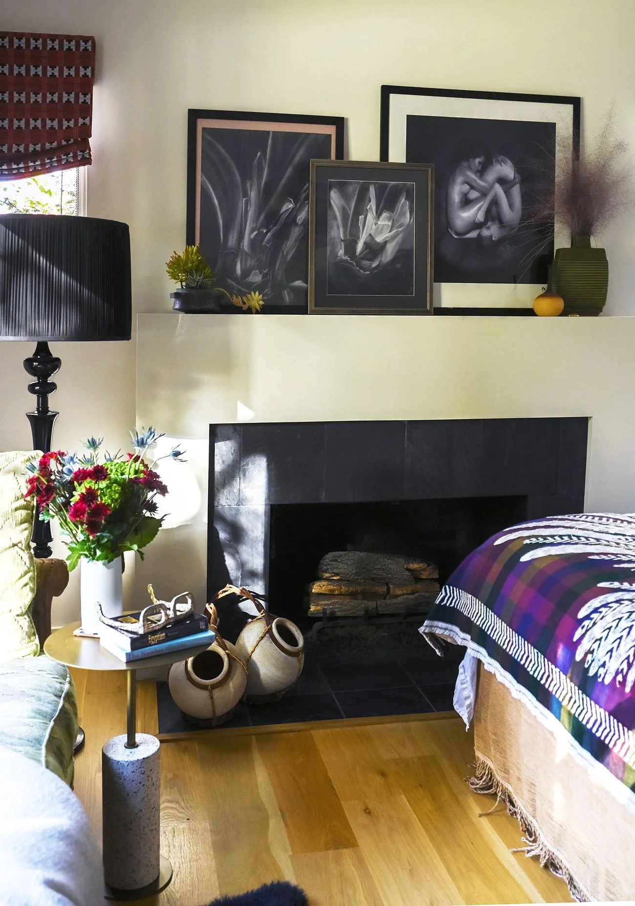 Living room corner with a black fireplace, framed artwork, side table with flowers and books, and a bed partially visible.