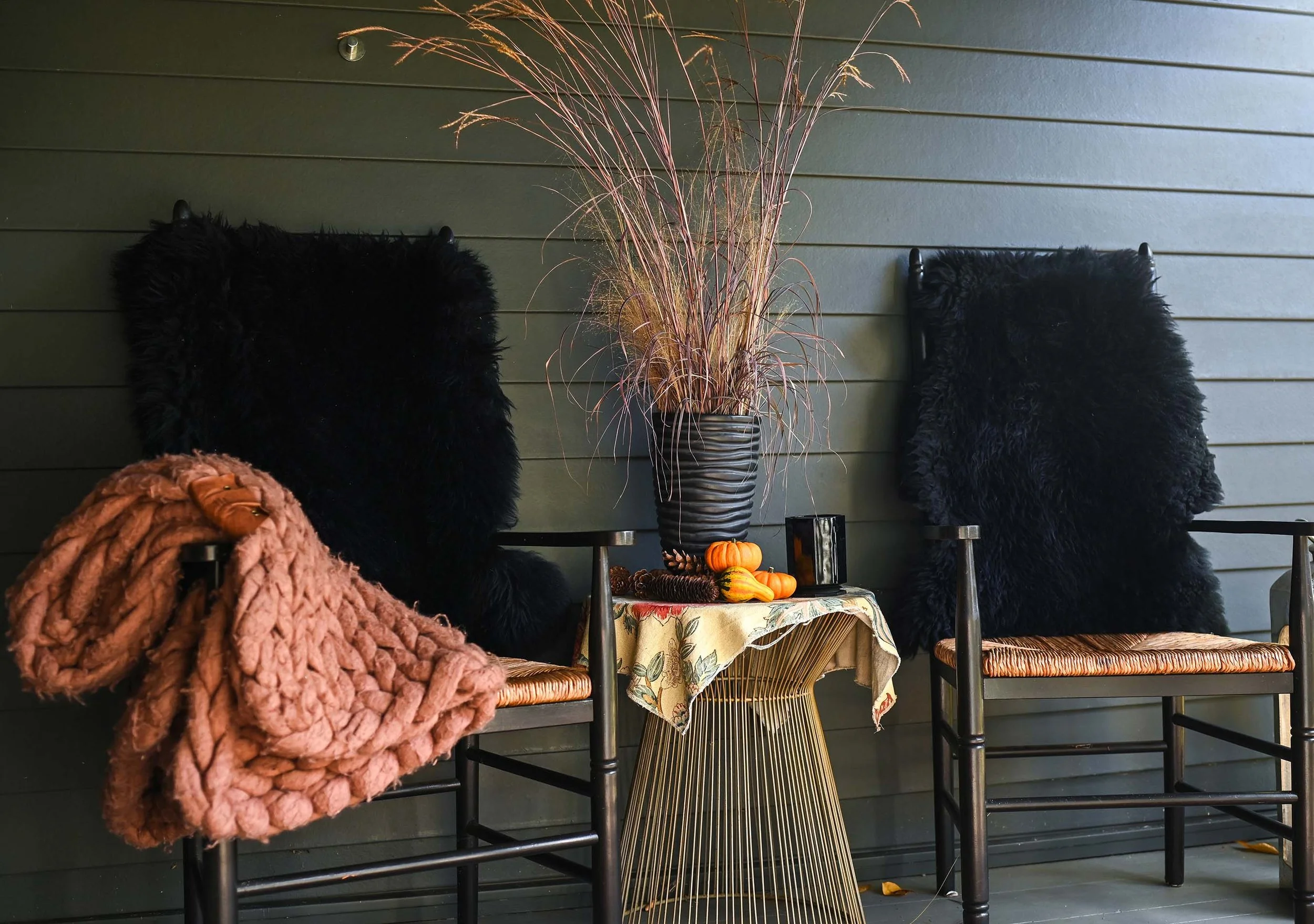 Two chairs with faux fur cushions on a porch beside a table with fall decorations and a tall plant.