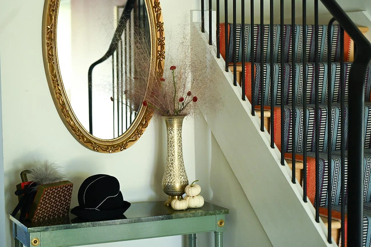 A gold-framed mirror hanging on a wall above a green console table. The table has decorative items including a large ornate vase with dried flowers and a small pumpkin. Part of a staircase with patterned carpeted steps and black metal railing is visi