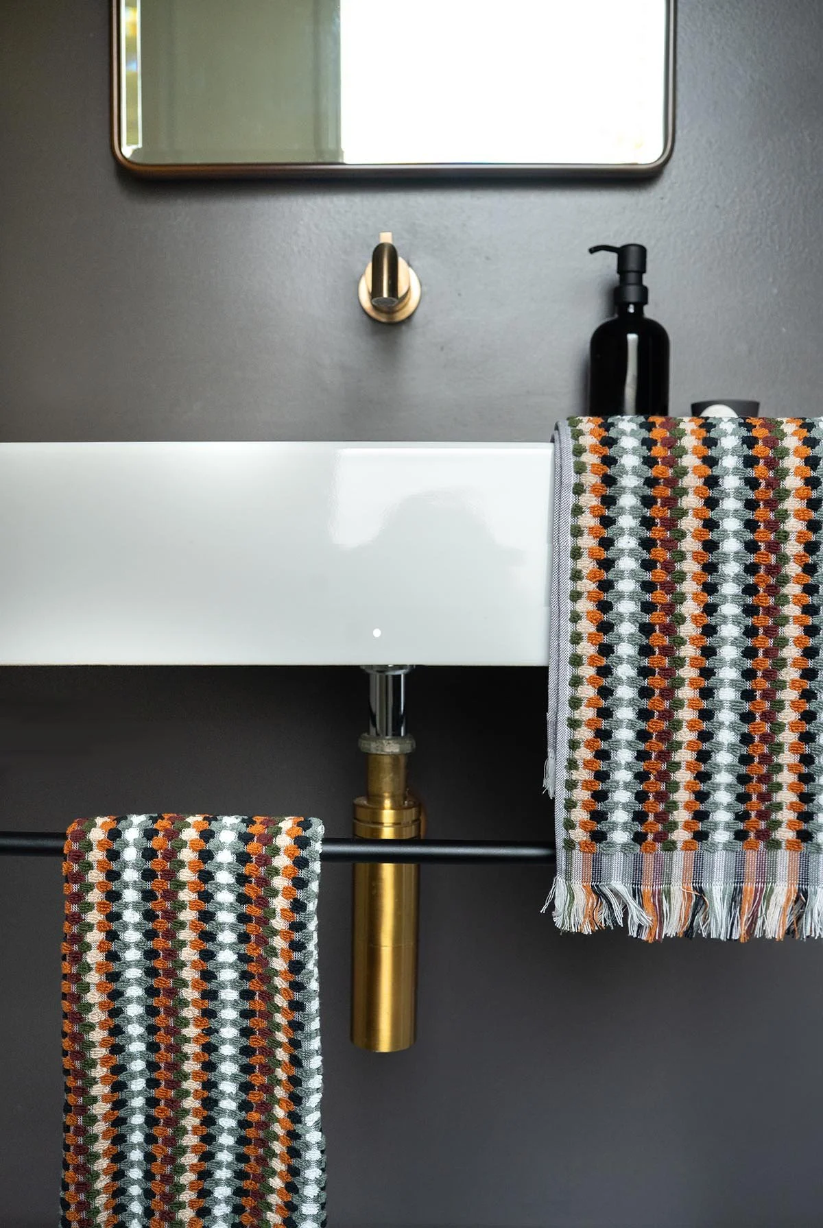 Bathroom sink with a black soap dispenser, a patterned towel hanging over the sink, a mirror above, and a black wall behind with a gold pipe visible beneath the sink.