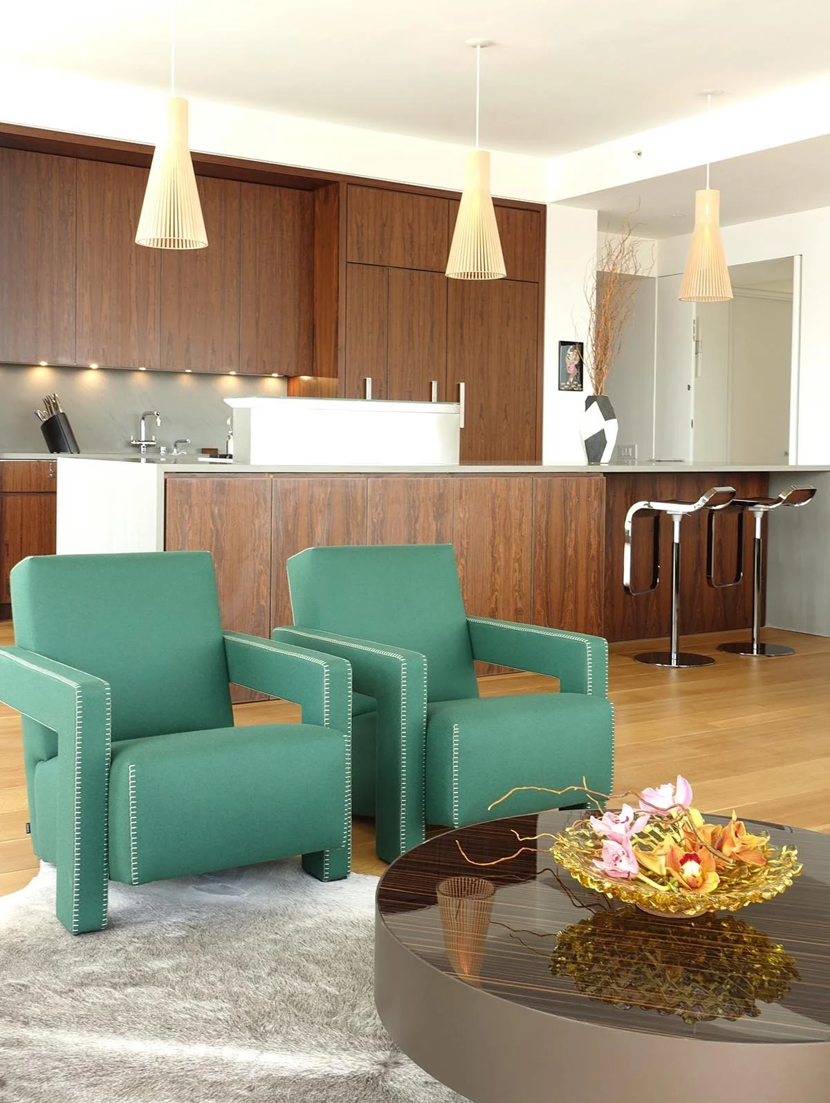 Living room with two green armchairs, a coffee table with a floral arrangement, and a kitchen area with wooden cabinets and a marble counter, hanging pendant lights, and two barstools.