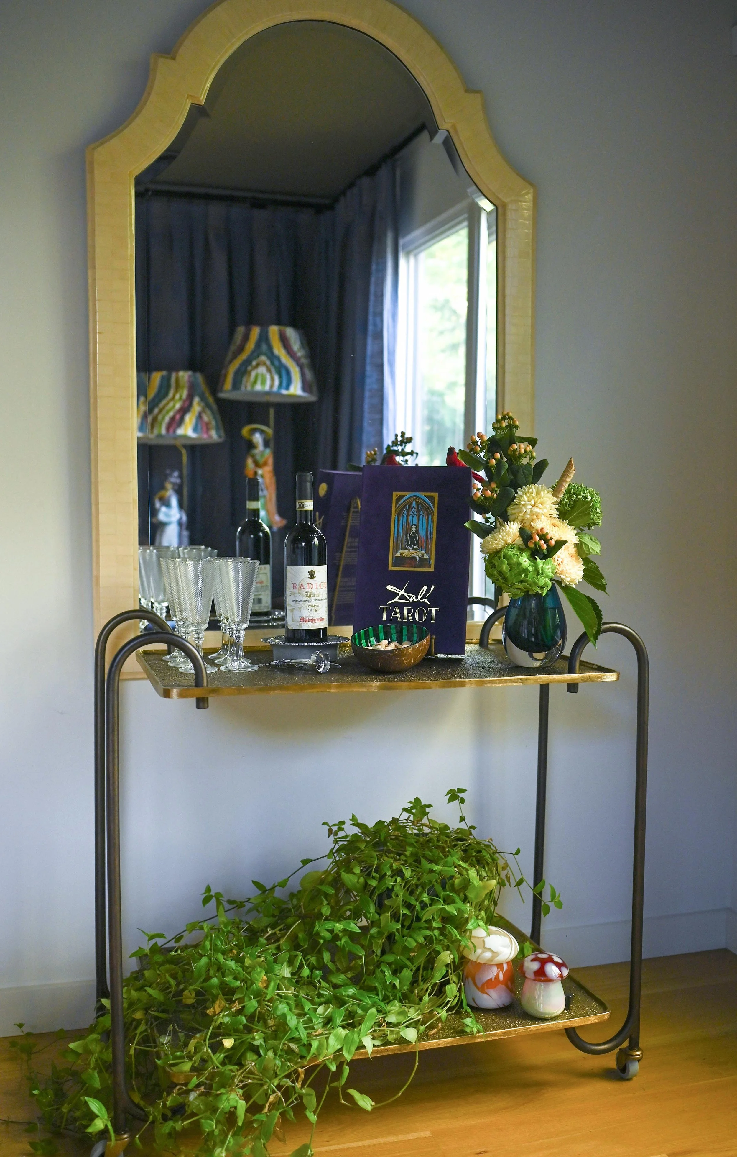 A decorative bar cart with wine bottles, glasses, a tarot card box, a blue vase with flowers, and a small bowl. A large mirror hangs on the wall behind the cart, reflecting lamps and a window with curtains. Green plants and decorative mushrooms are o