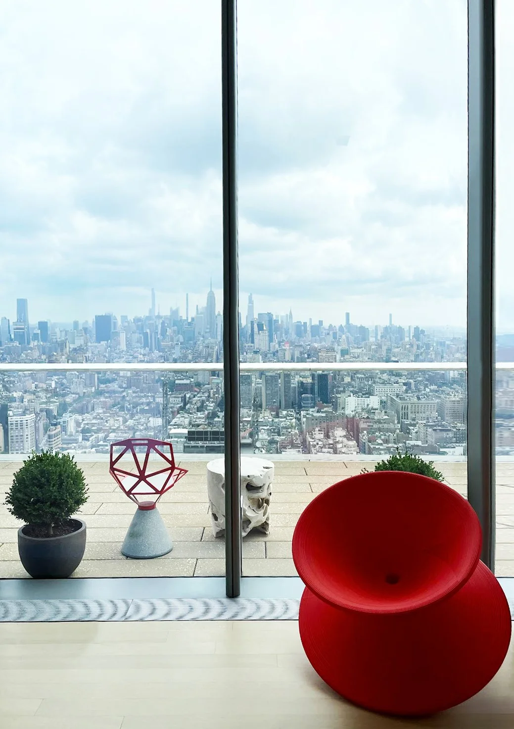 Interior view from a high-rise building with a red modern chair, two potted plants, a white sculpture, and a large glass window showing a city skyline with tall skyscrapers, including the Empire State Building.