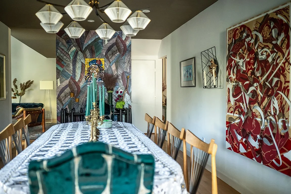 A dining room with a long table covered with a patterned tablecloth, surrounded by wooden chairs. The table has a gold candelabra with teal candles and a small decorative tray. On the wall at the end, there is a colorful abstract mural and a small Ti