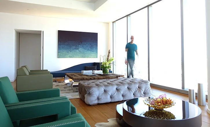 Modern living room with green armchairs, a tufted gray ottoman, a coffee table with a flower arrangement, and a man walking past large floor-to-ceiling windows.