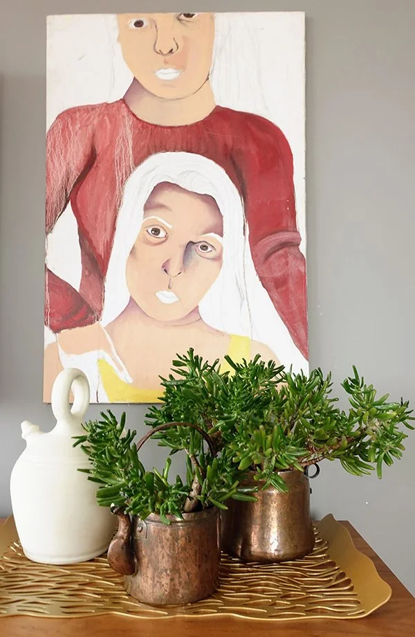 A still life of a white ceramic vase, two copper pots with green succulent plants, placed on a wooden surface with a gold decorative tray. In the background, a multicolored portrait painting of two women, one with white hair and the other with a partial view, wearing red and yellow clothing, hangs on a gray wall.