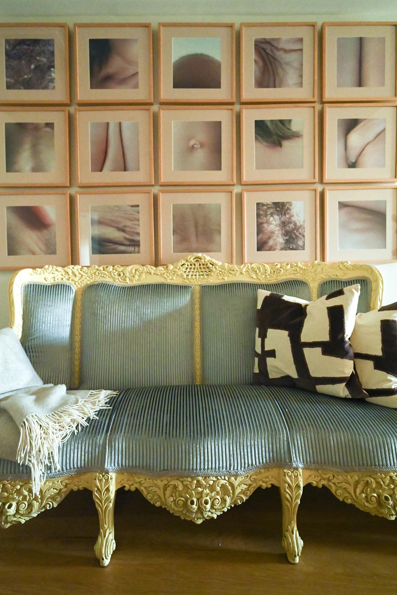 A vintage green velvet sofa with ornate gold woodwork detail, placed in front of a wall decor consisting of a grid of pink-framed photographs or artworks, with three black and white patterned throw pillows and a fringed white throw blanket.