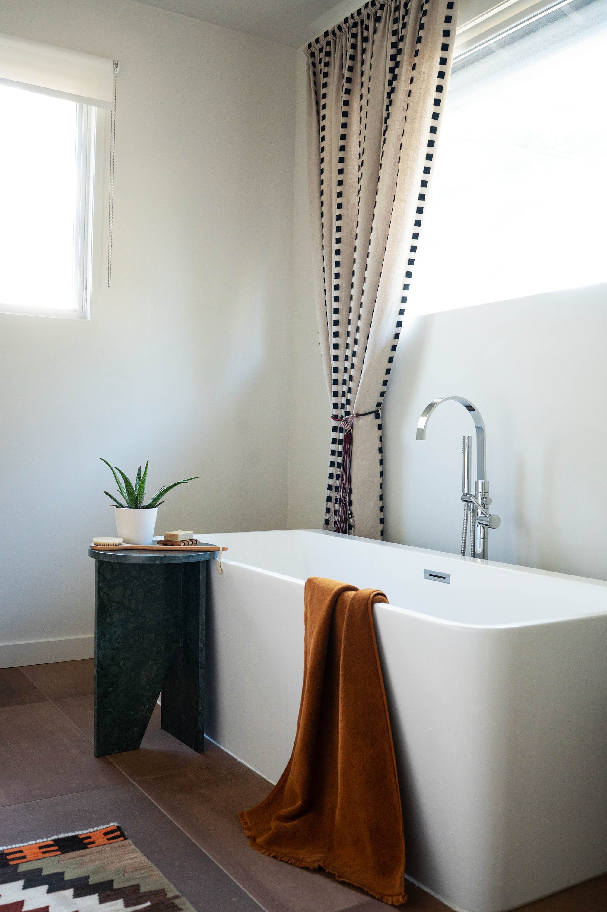 A modern bathroom with a white bathtub next to a window with black and white patterned curtains, a small dark green marble side table holding a potted plant, a soap dish, and a brush, and a brown bath towel hanging over the edge of the tub.