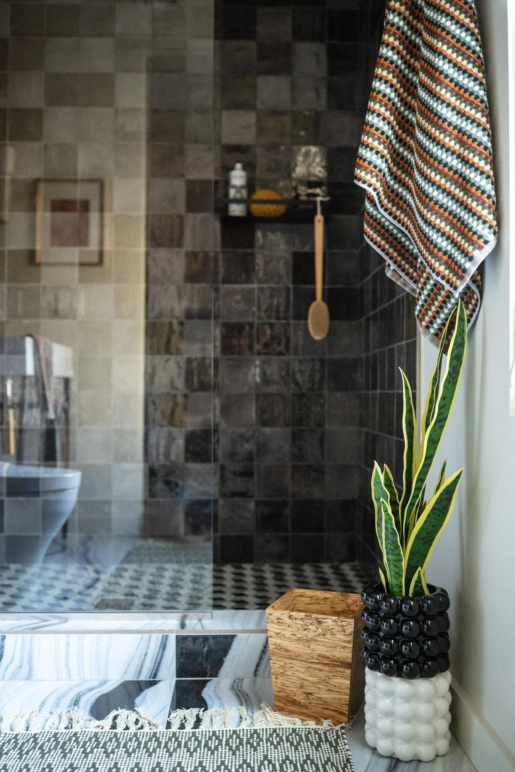 Decorative bathroom corner with a snake plant in a black-and-white beaded vase, a wooden box, a patterned fabric, and a multicolored towel hanging near the shower.
