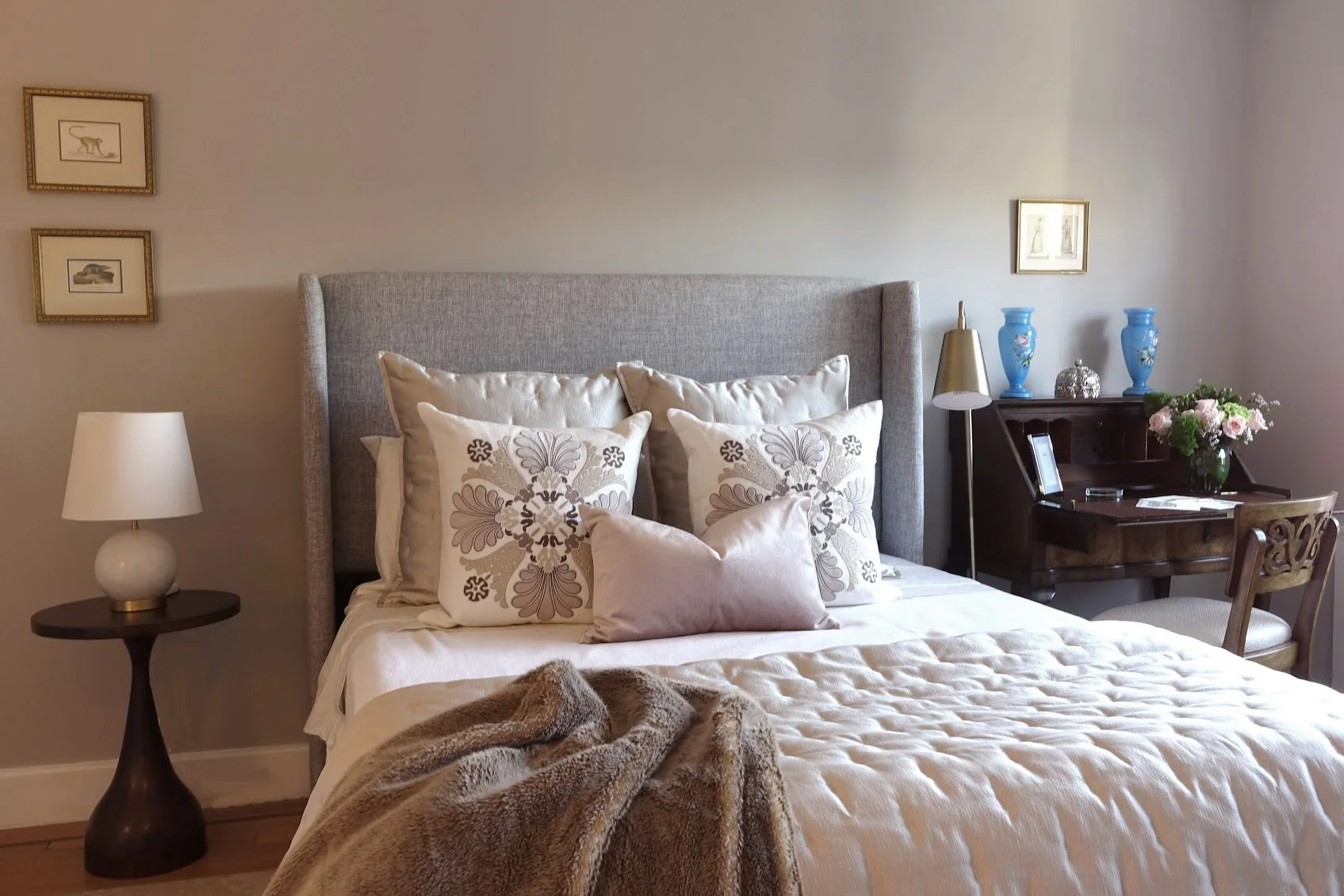 A neatly made bed with decorative pillows and a furry blanket, in a cozy bedroom with a bedside lamp, framed artwork, and a small desk with vases, flowers, and a chair.