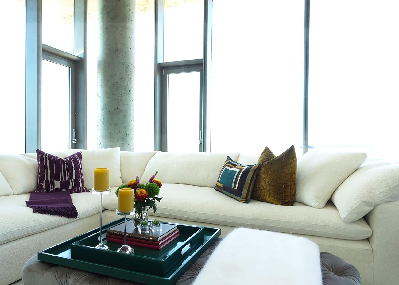 Modern living room with white sectional sofa, decorative pillows, a coffee table with yellow candles and a flower arrangement, large windows with natural light.