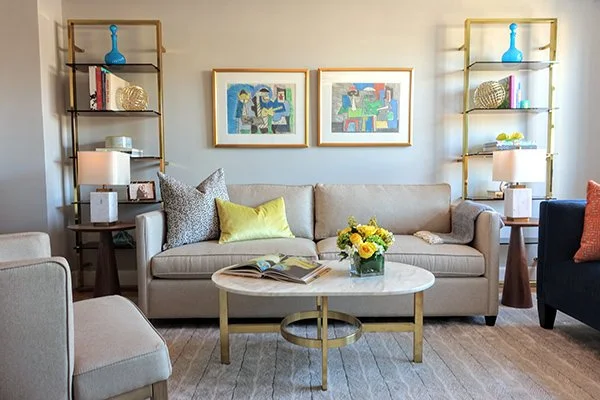 Living room with a beige sofa adorned with colorful pillows, a white marble coffee table with a flower arrangement, and two gold-framed shelving units with decorative items and books. Two framed modern art pieces hang above the sofa.