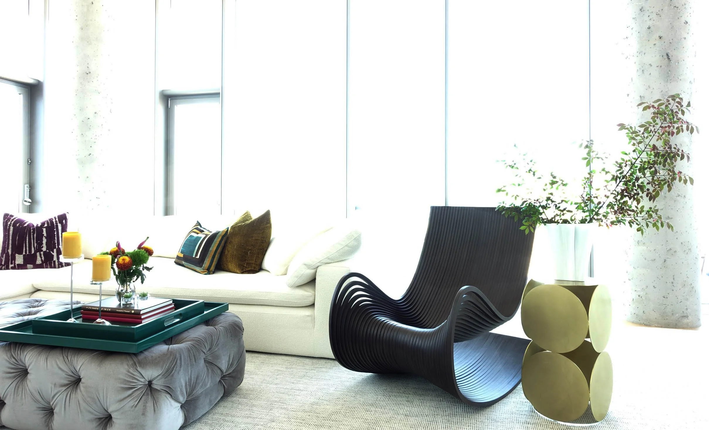 Modern living room with a white sofa, decorative pillows, a black wavy lounge chair, a green side table with a plant, windows letting in natural light, and a gray tufted ottoman with tray and candles.