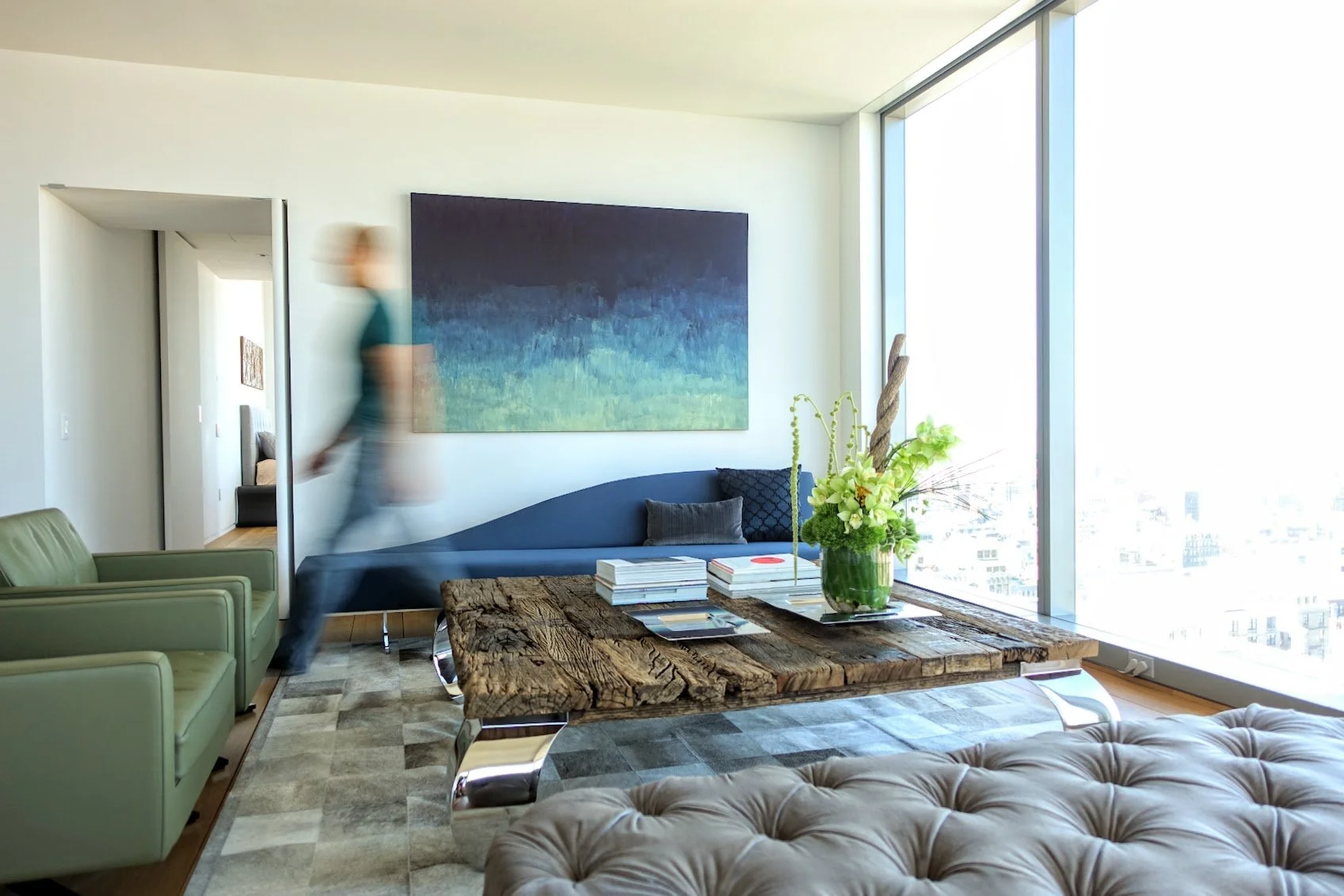 Modern living room with large window, wooden coffee table with flowers, green arm chairs, abstract painting, person blurred walking through, gray tufted chaise lounge, patterned rug.