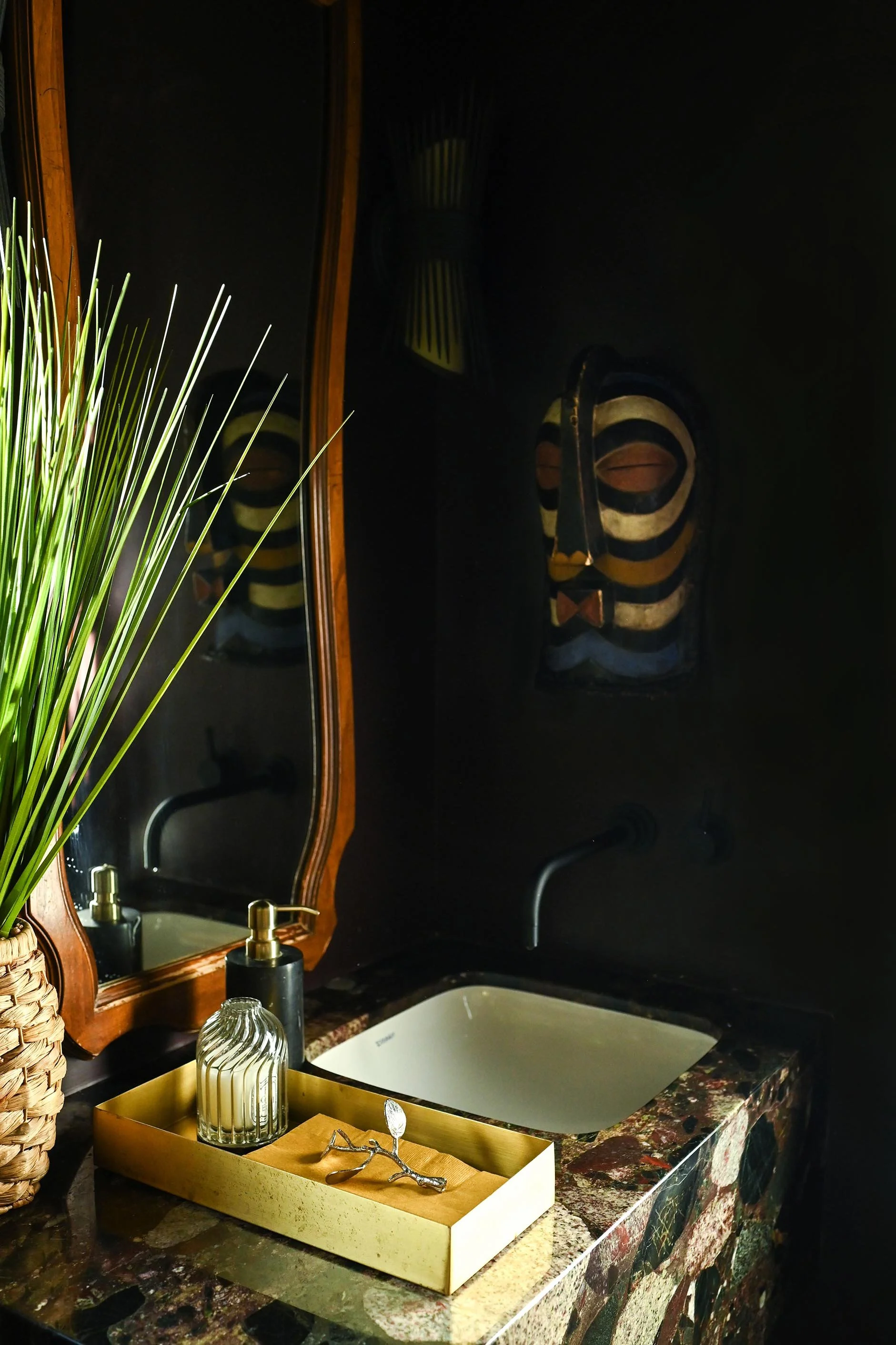 Bathroom sink with gold soap dispenser, candle, and decorative tray on marble countertop, dark wall with African mask and mirror reflecting sink