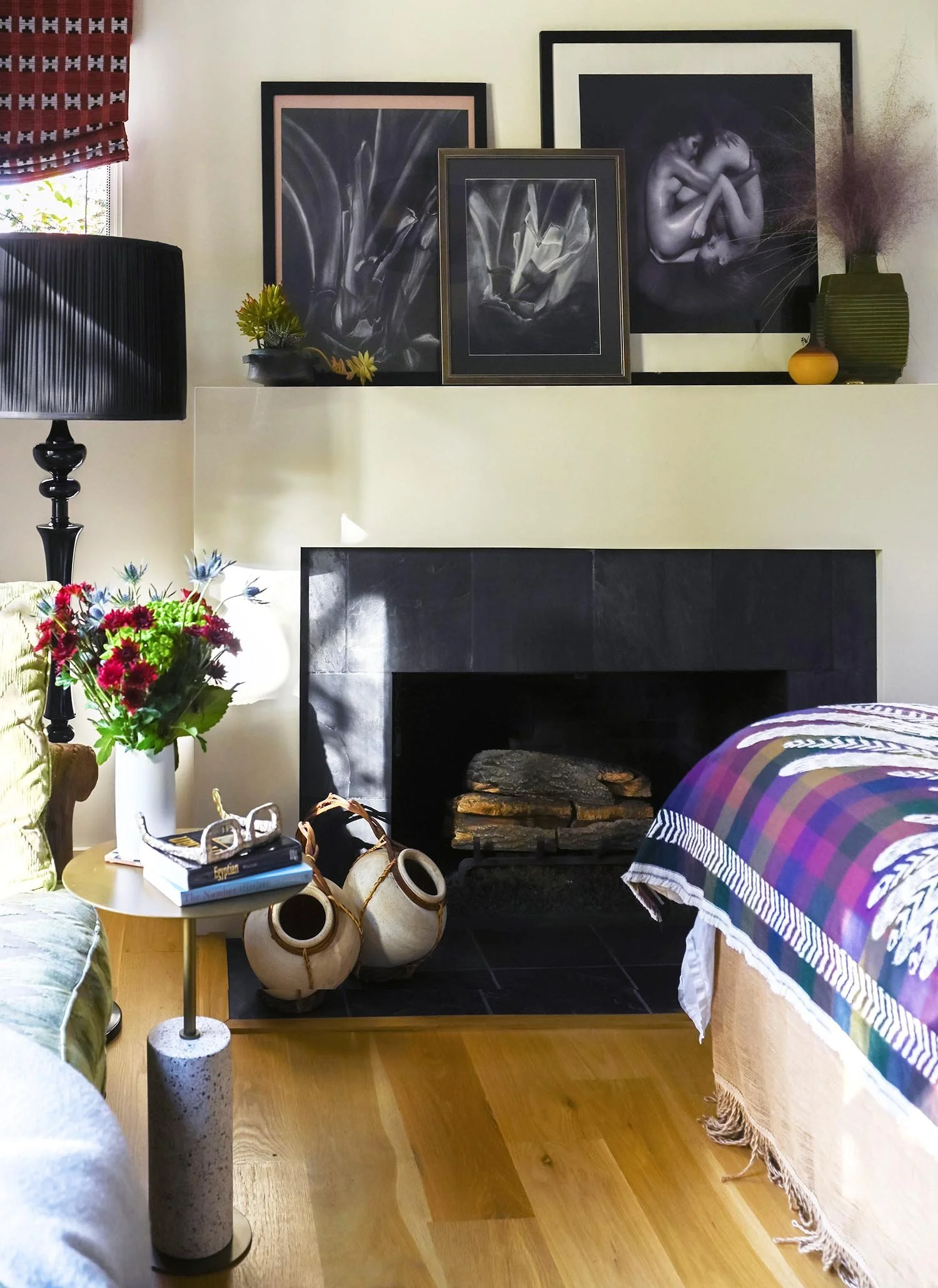 Cozy living room corner with a black fireplace, colorful flowers on a small side table, framed art on a wall above the fireplace, and a bed with a multicolored quilt.