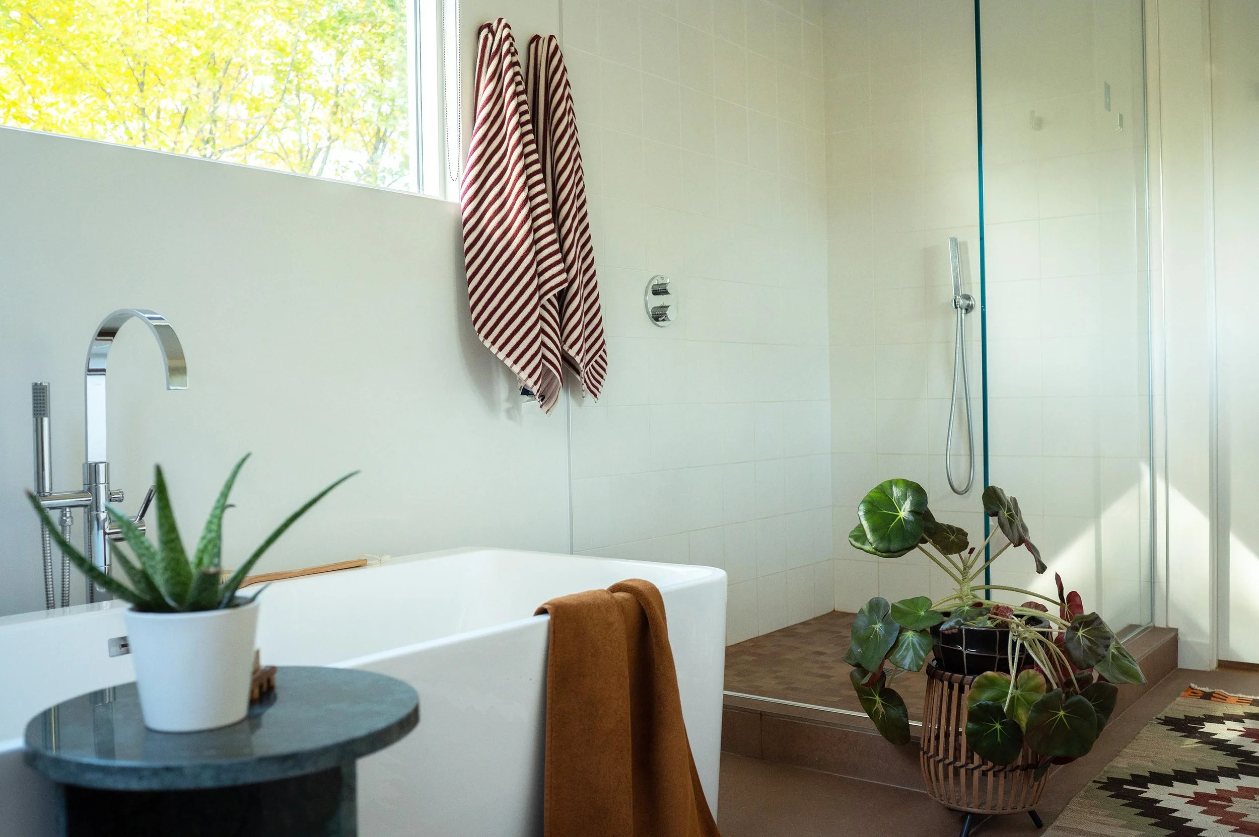 A modern bathroom featuring a white bathtub with a brown towel draped over it, a potted aloe plant on a small table, a hanging towel with red and white stripes, a large window showing trees outside, a walk-in shower with a glass door, a potted plant 