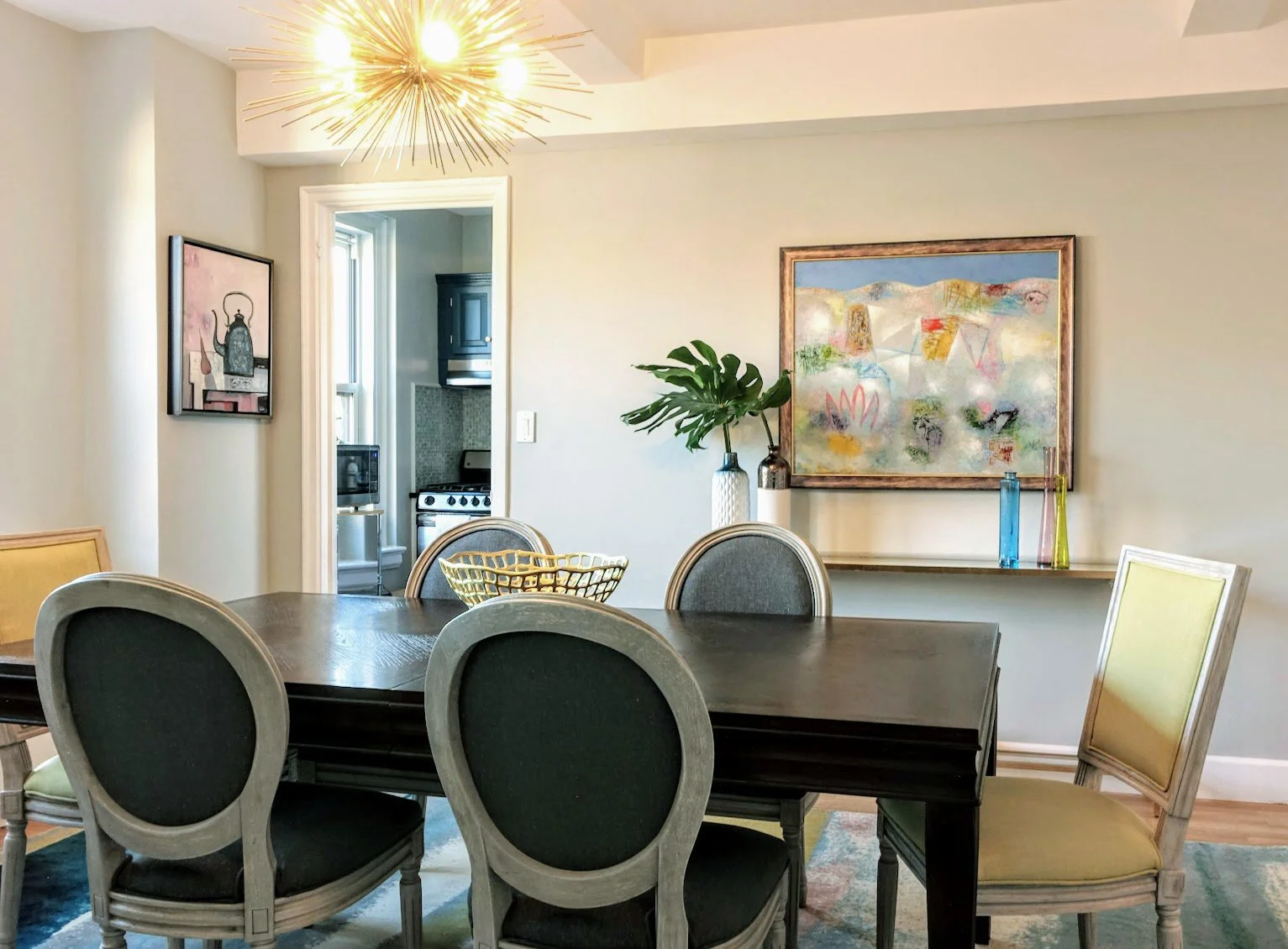 A dining room featuring a dark wood table surrounded by six upholstered chairs with gray and yellow designs. A modern gold chandelier hangs from the ceiling. The wall has a large abstract painting, and a small shelf with decorative glass vases and a 