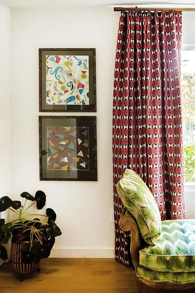 Living room corner with two framed abstract art pieces on white wall, a window with red patterned curtains, a green velvet armchair, and a potted plant on the wooden floor.