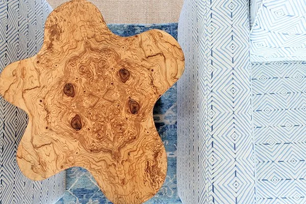 A uniquely shaped wooden side table with a natural wood finish, placed next to a patterned sofa or bed.